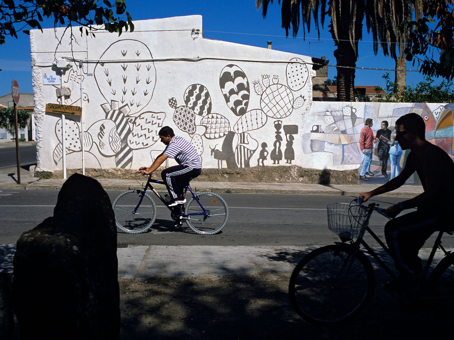 Two cyclists ride past a bold black and white street mural on a sunny urban street corner.