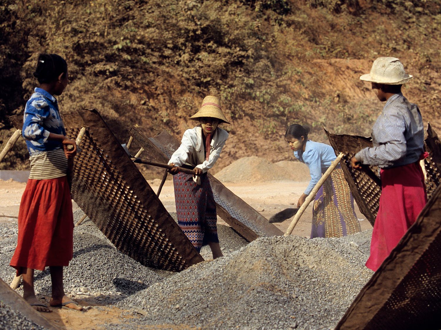 Women in traditional attire sifting gravel with mesh screens at an outdoor construction or mining site.