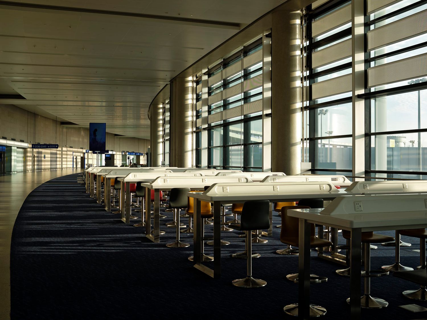 Row of white tables with bar stools in an empty modern airport terminal with large windows and blue carpet.
