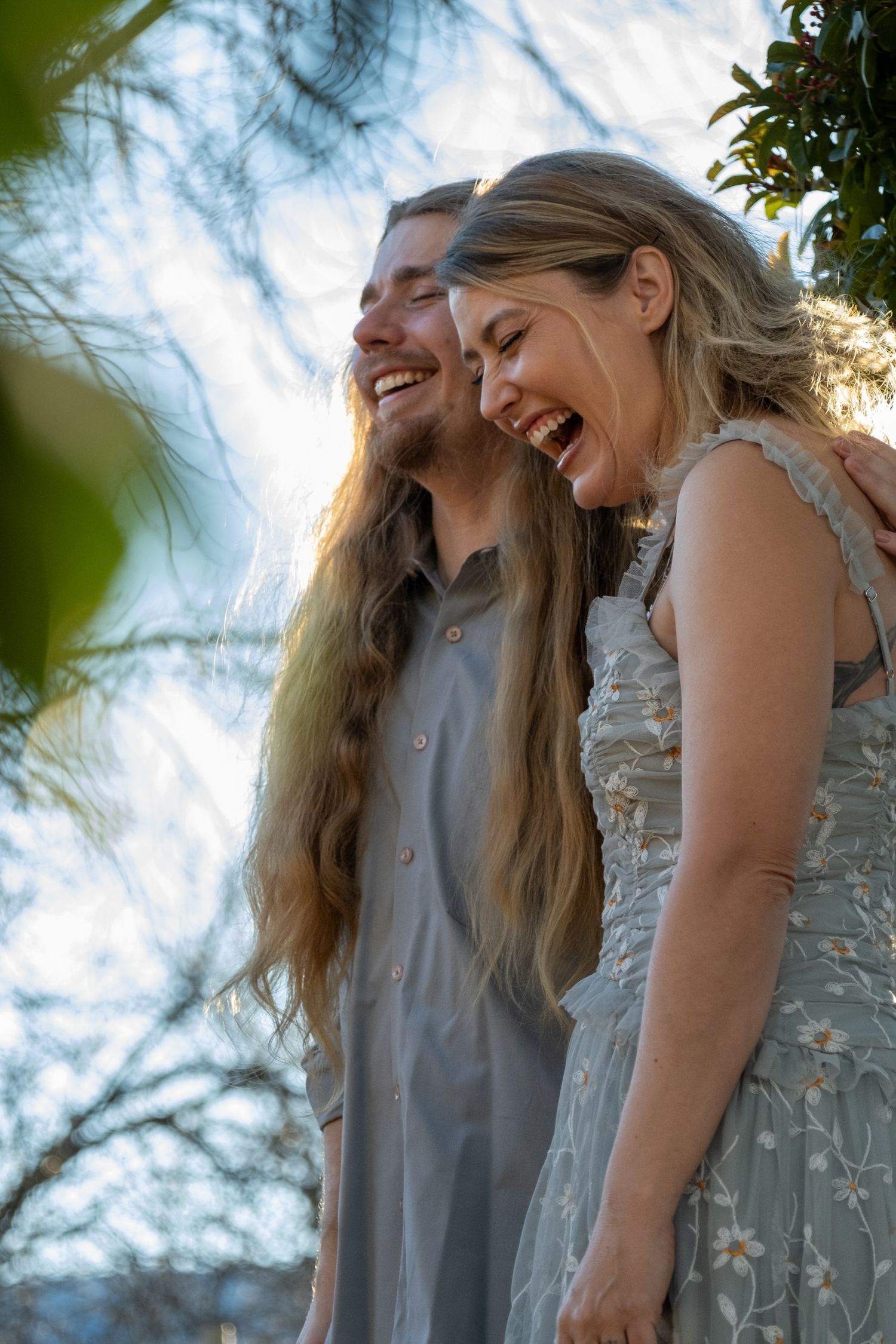 Happy laughing couple with long hair embracing outdoors in soft natural light among green foliage.