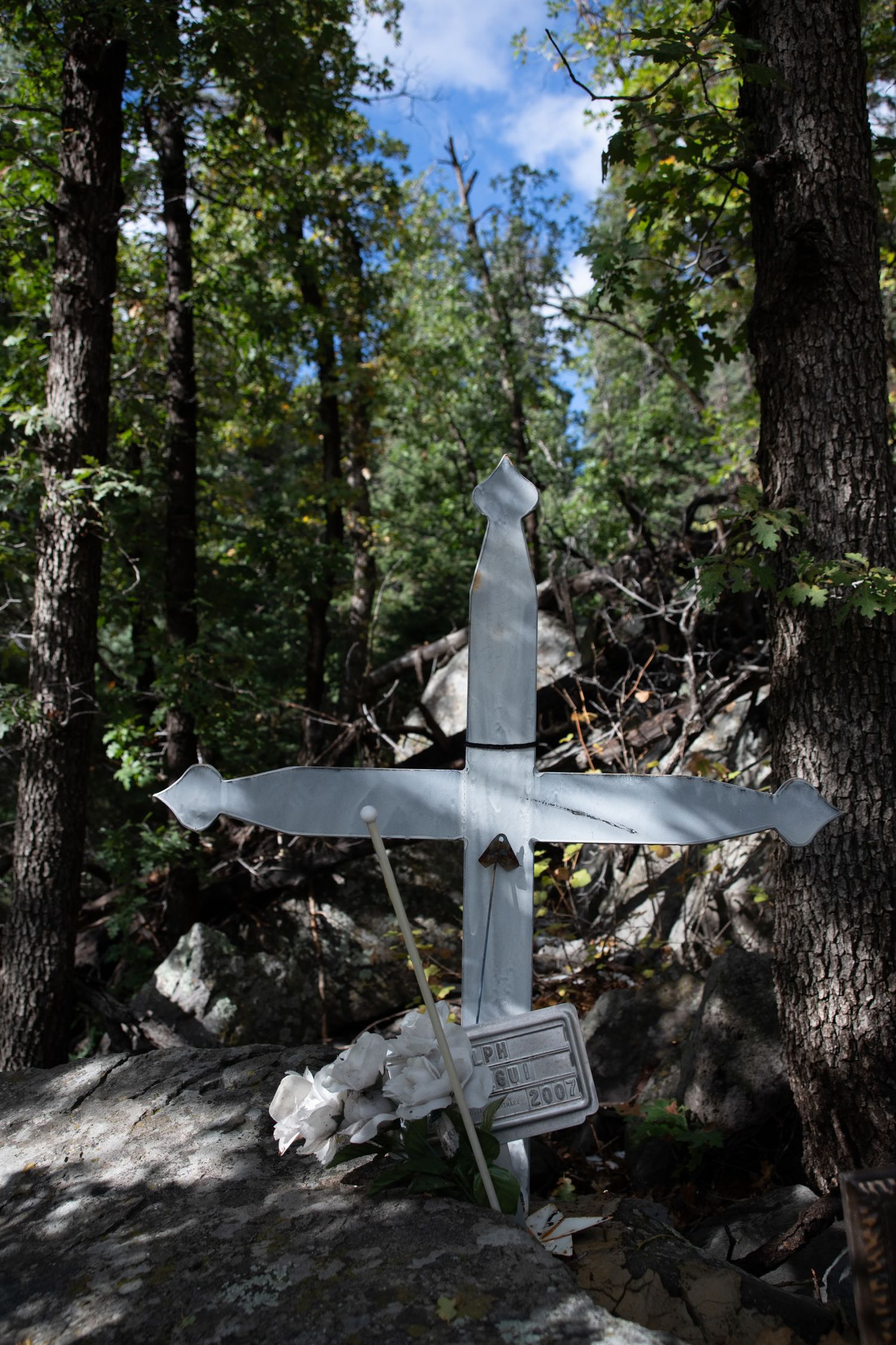White metal memorial cross with plaque surrounded by trees, rocks, and white flowers in forest setting.