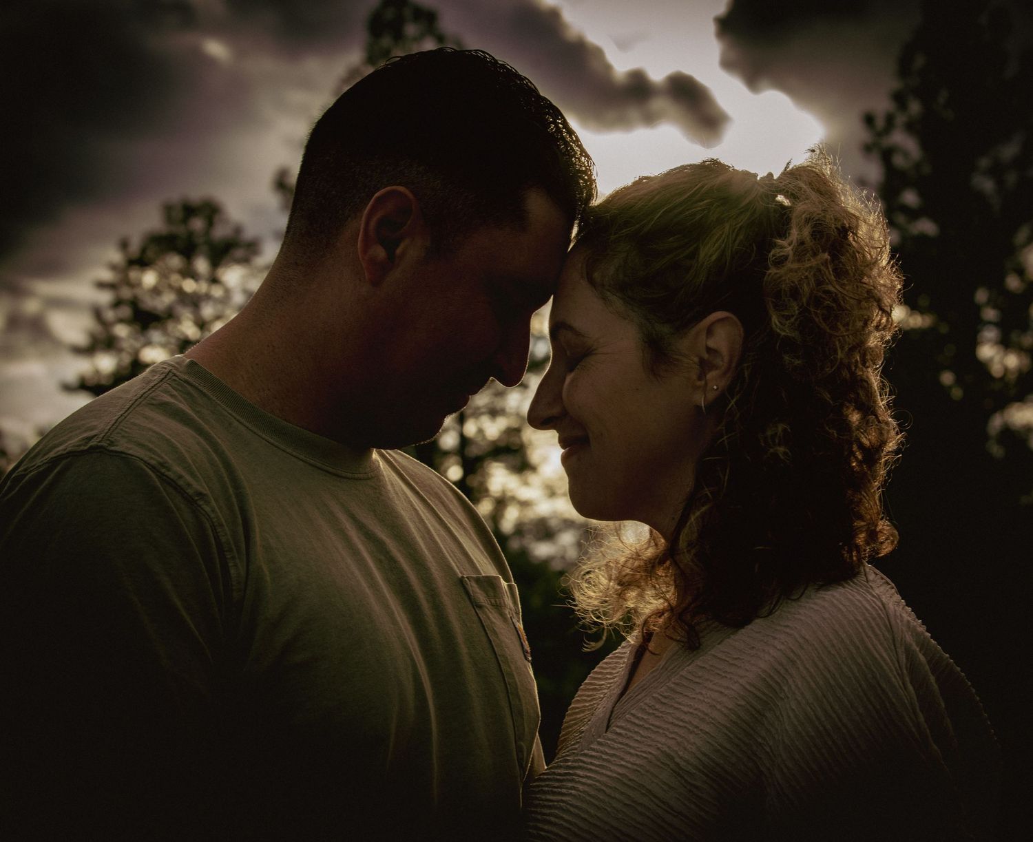 Romantic couple touching foreheads under dramatic cloudy sky with moody dark lighting outdoors.