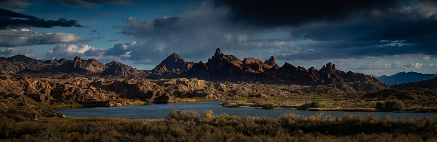 Dramatic panoramic view of rugged desert mountains reflected in a calm lake under stormy skies.