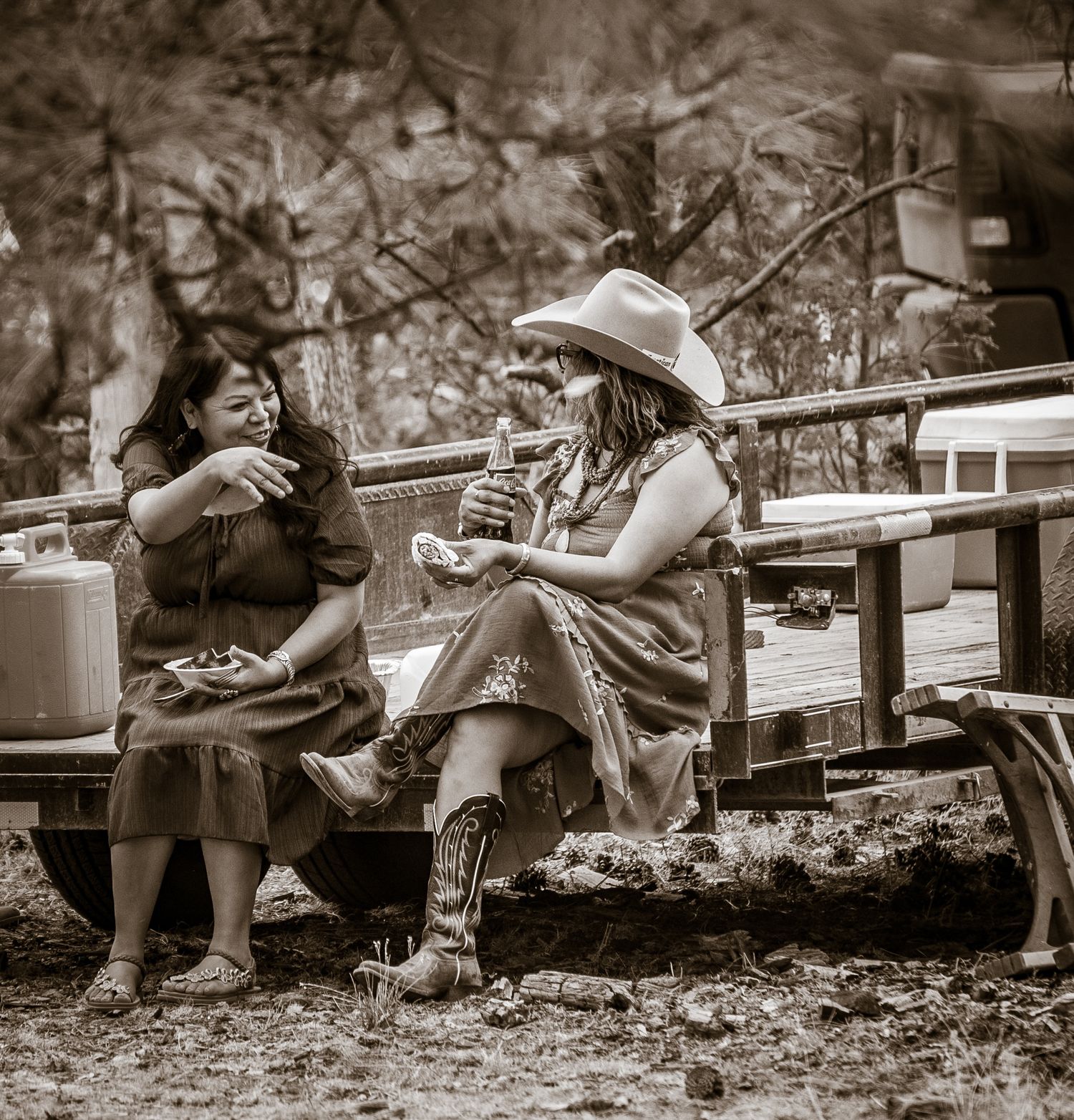 Two women in western attire sitting and chatting by a wagon in sepia-toned photograph.