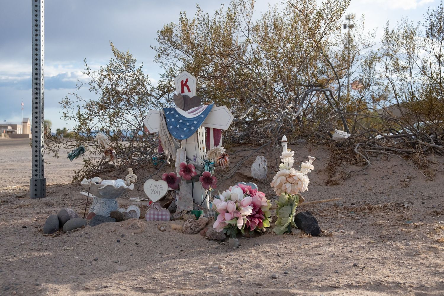Roadside memorial with letter K heart cross and colorful flowers in sandy desert.