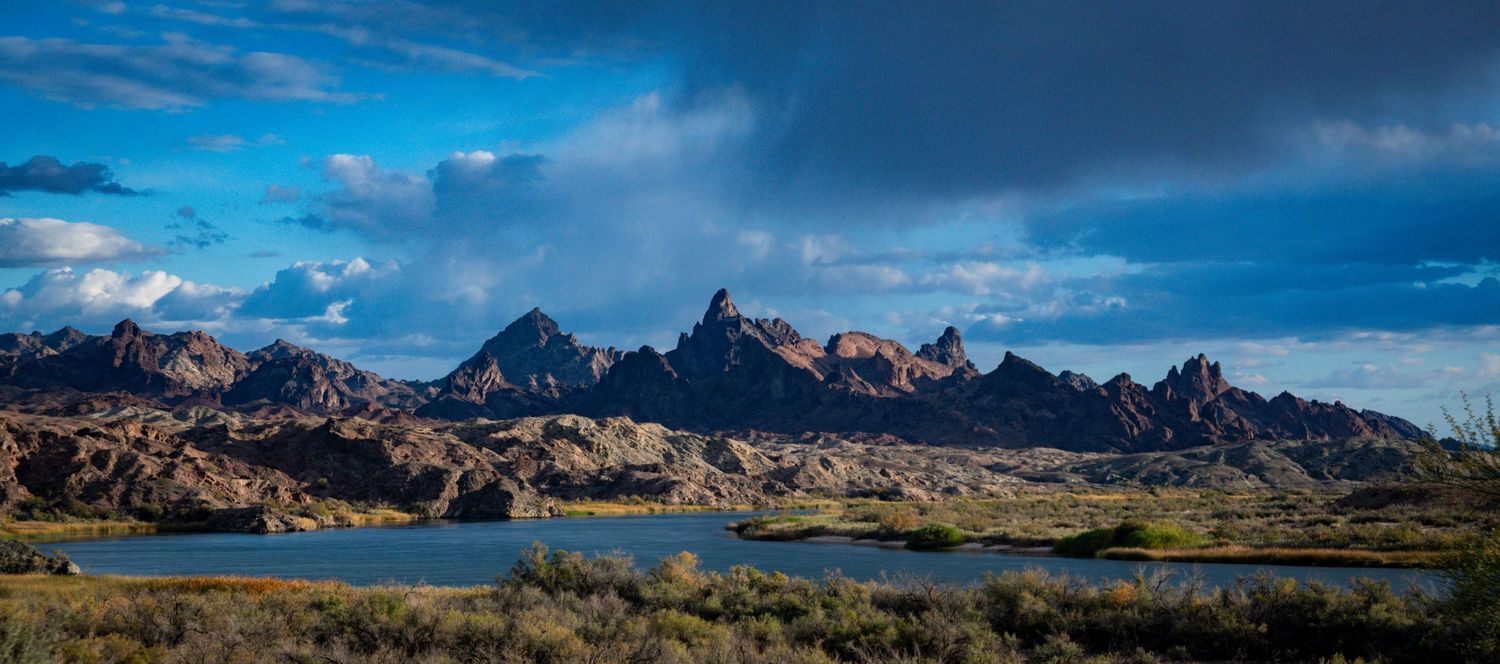 Rocky mountain landscape with blue lake reflecting dramatic storm clouds in arid desert terrain.