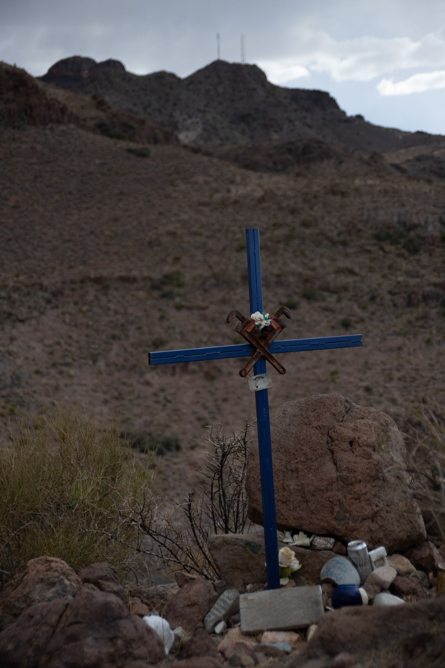 Blue memorial cross with flowers and stones at base set against a rugged desert mountain landscape.