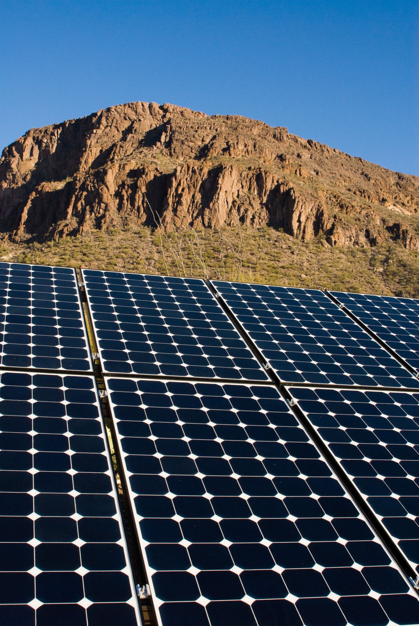 Solar panels array in Arizona desert with dramatic red rock mountain backdrop under clear blue sky.