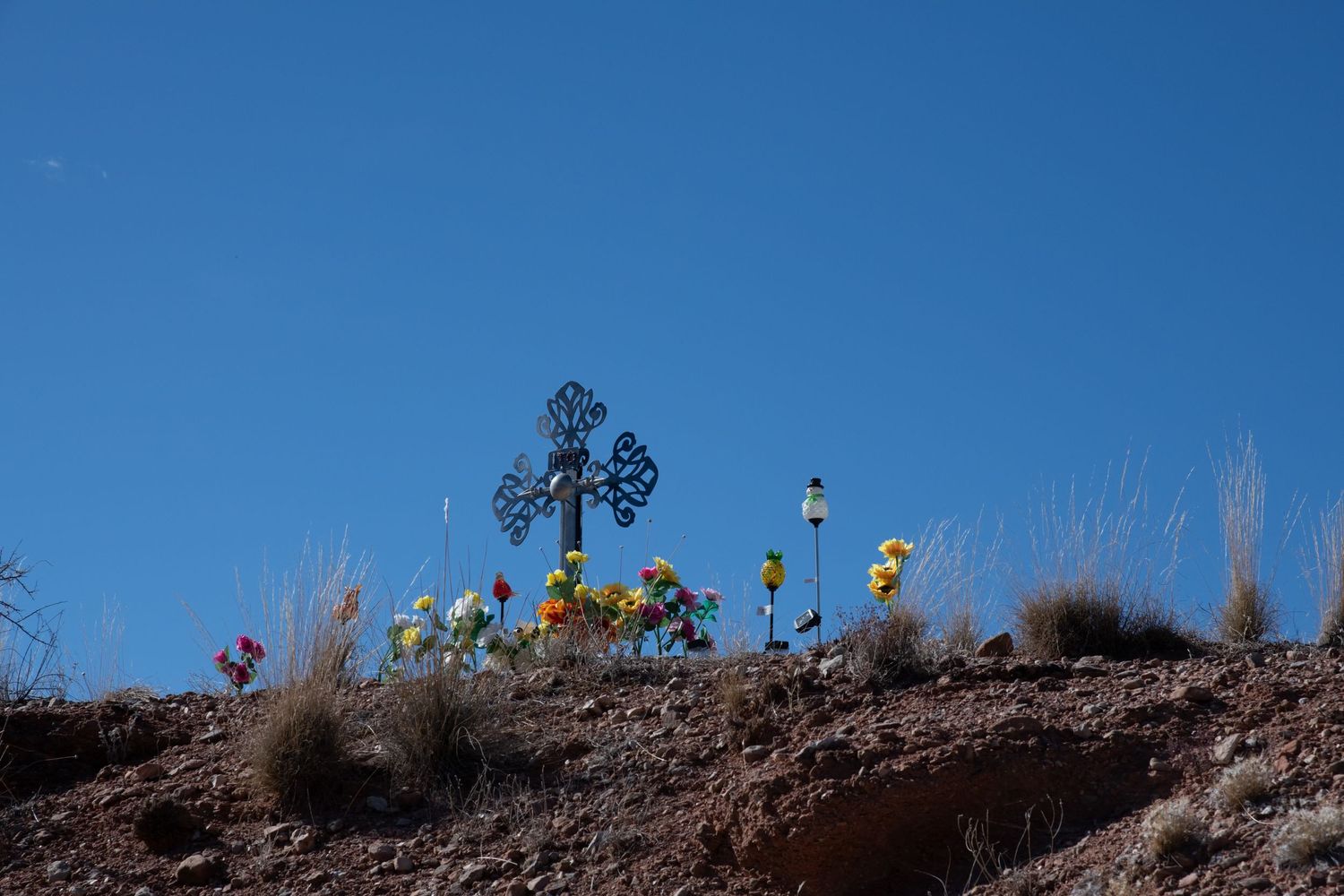 Decorative metal cross memorial with colorful artificial flowers on a rocky hillside against blue sky.