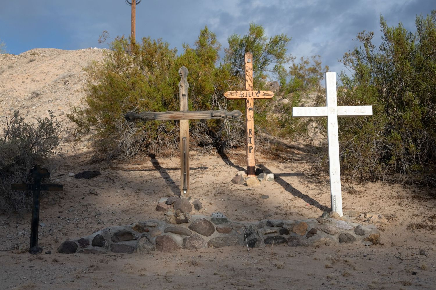 Three memorial crosses in desert sand with rocks, surrounded by green desert shrubs.