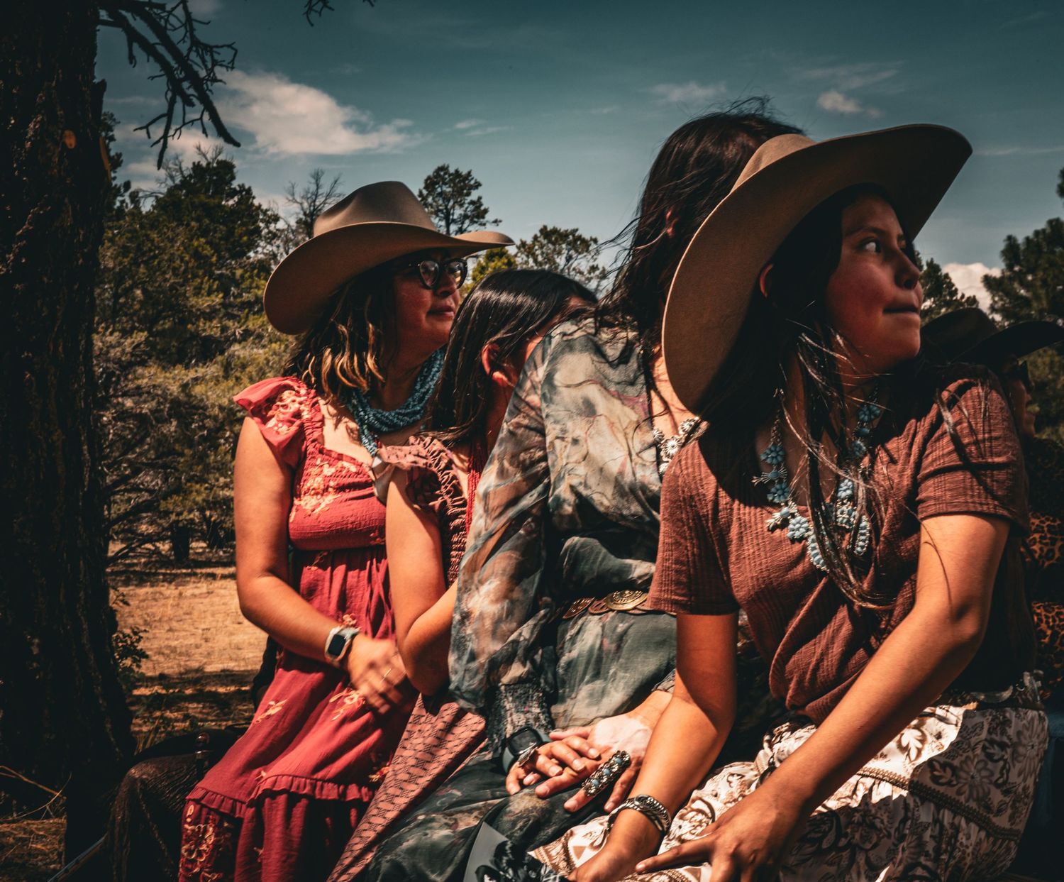 Three women wearing wide-brim hats and boho dresses sit outdoors under a blue sky in a rustic desert setting.