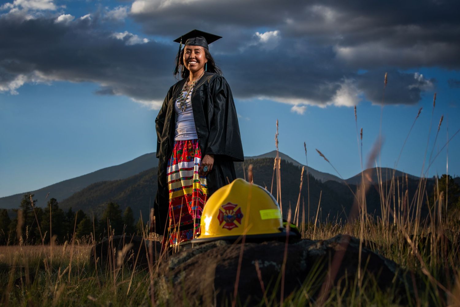 Smiling graduate in cap and gown stands in a mountain meadow with a yellow firefighter helmet nearby.