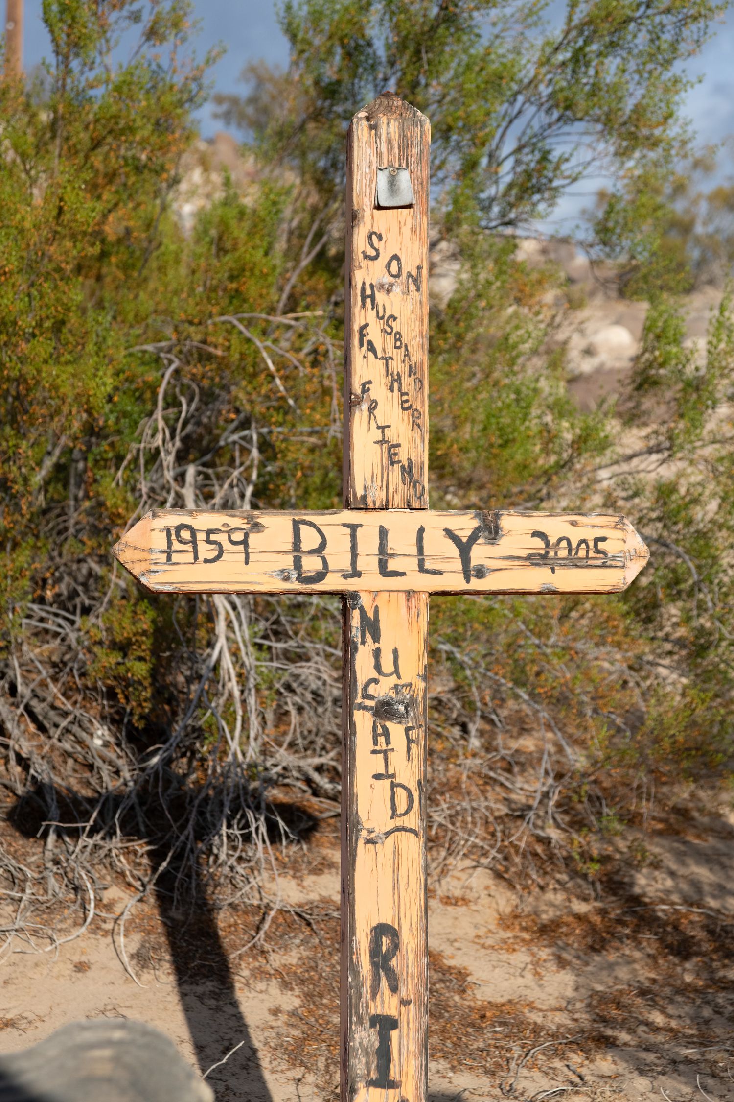 Weathered wooden memorial cross for Billy 1959-2005 with handwritten inscriptions in desert.