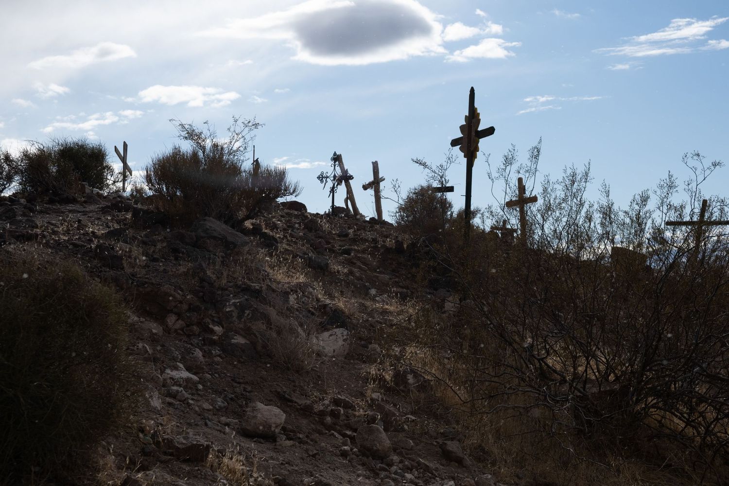 Silhouetted memorial crosses dot a rugged desert hillside against a dramatic blue sky with scattered clouds.