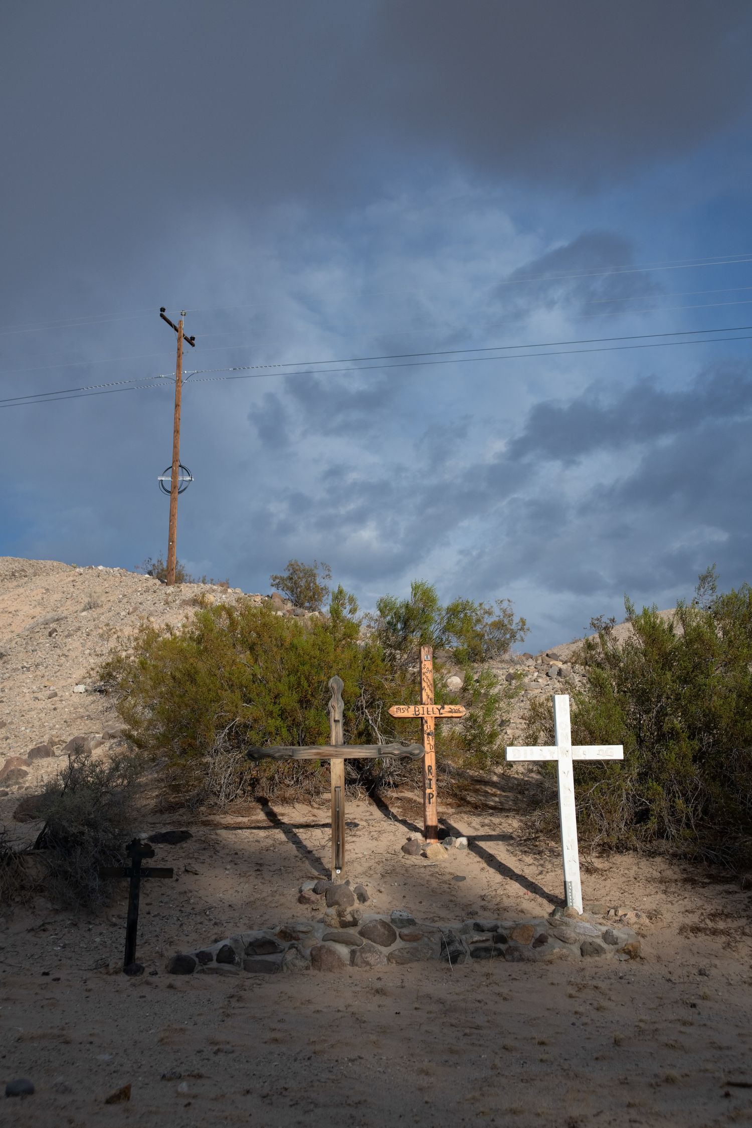Three roadside memorial crosses in desert sand beneath dramatic stormy sky with power pole.