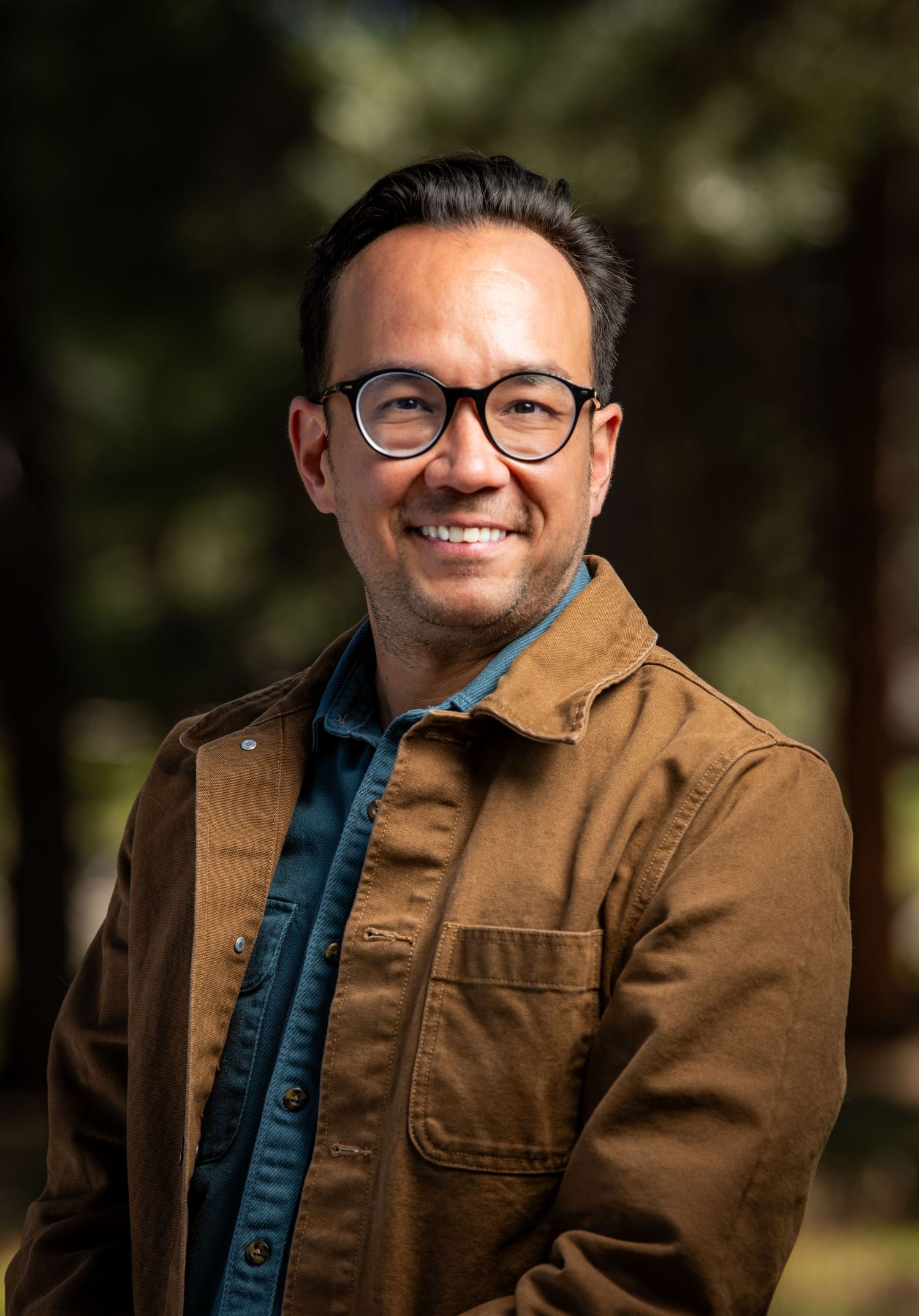 Smiling Asian man wearing glasses and brown canvas jacket posing outdoors among pine trees.