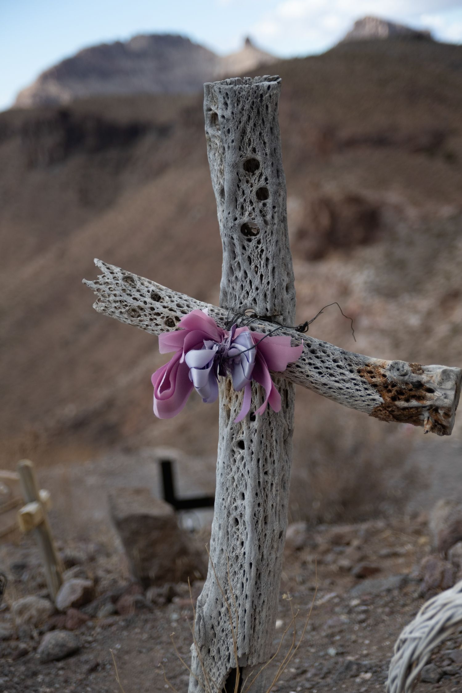 A weathered driftwood cross decorated with faded pink fabric flowers in a desert mountain cemetery.