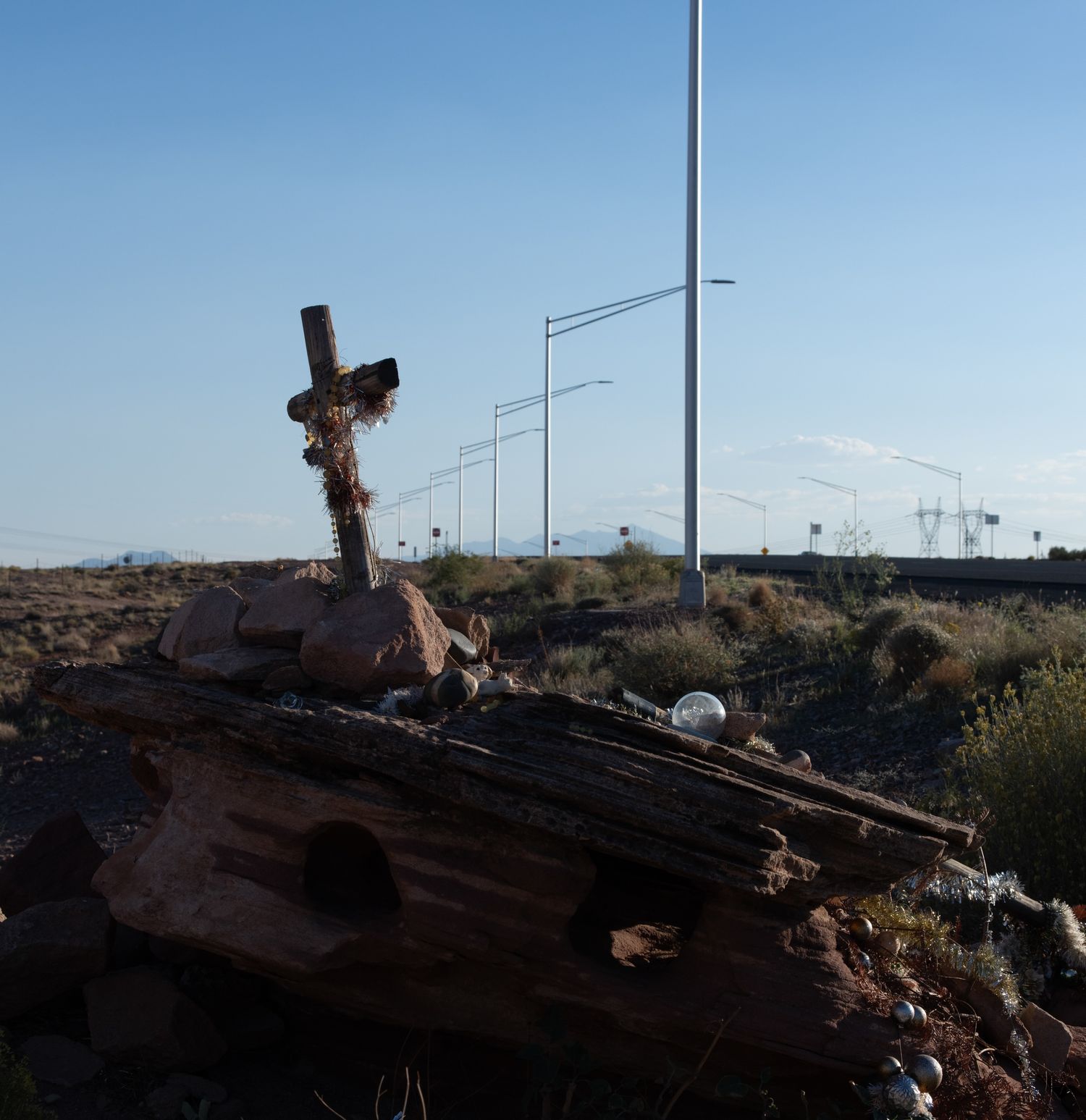 Weathered wooden memorial cross leaning among debris near highway light poles at dusk.