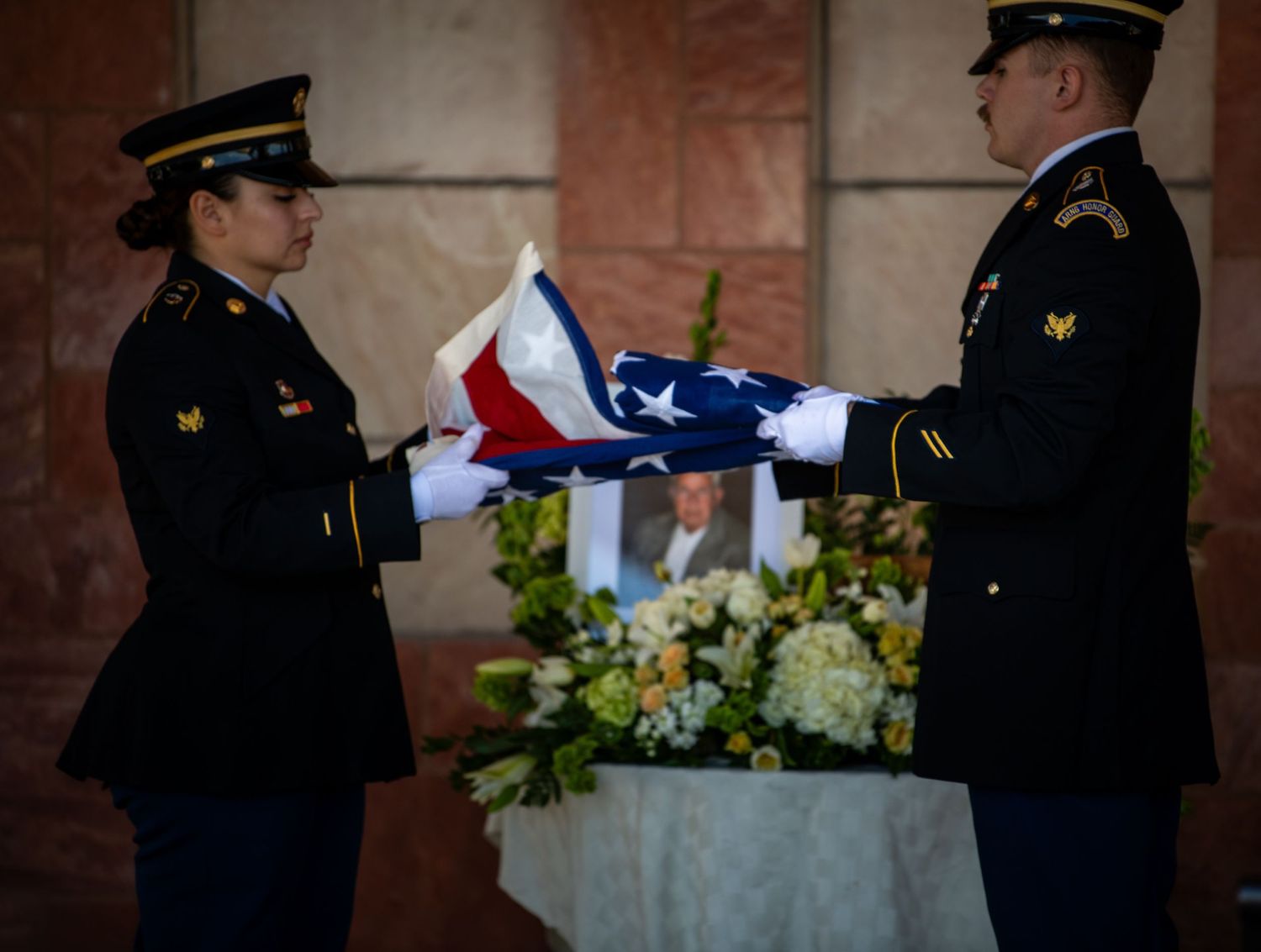 Two military soldiers in dress uniform folding an American flag at a memorial service with flowers.