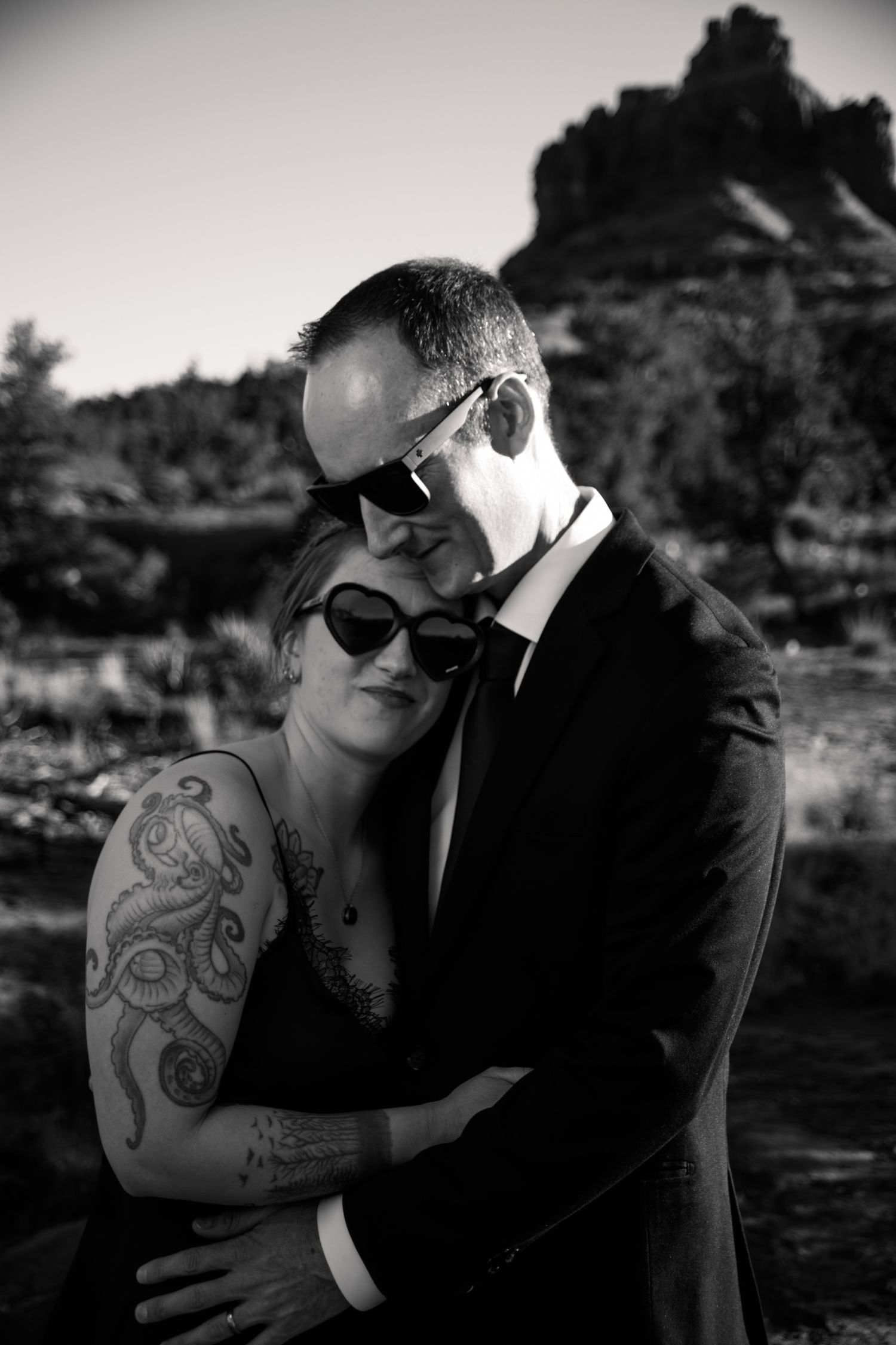 Black and white photo of a tattooed woman and suited man embracing near red rock formations.