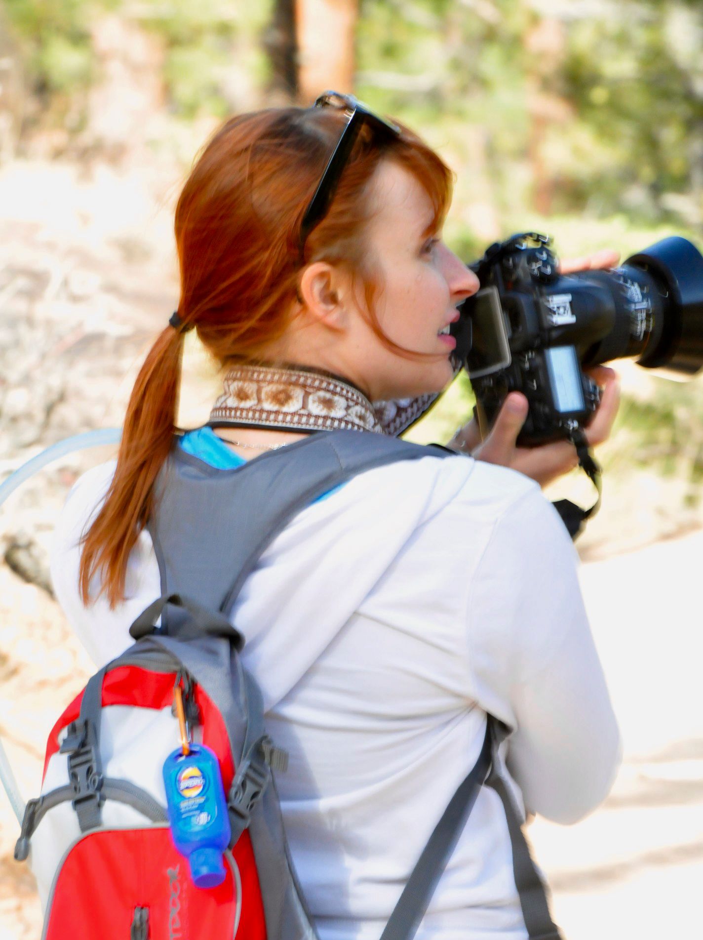 Red-haired female photographer shooting with DSLR camera and telephoto lens wearing red backpack outdoors.