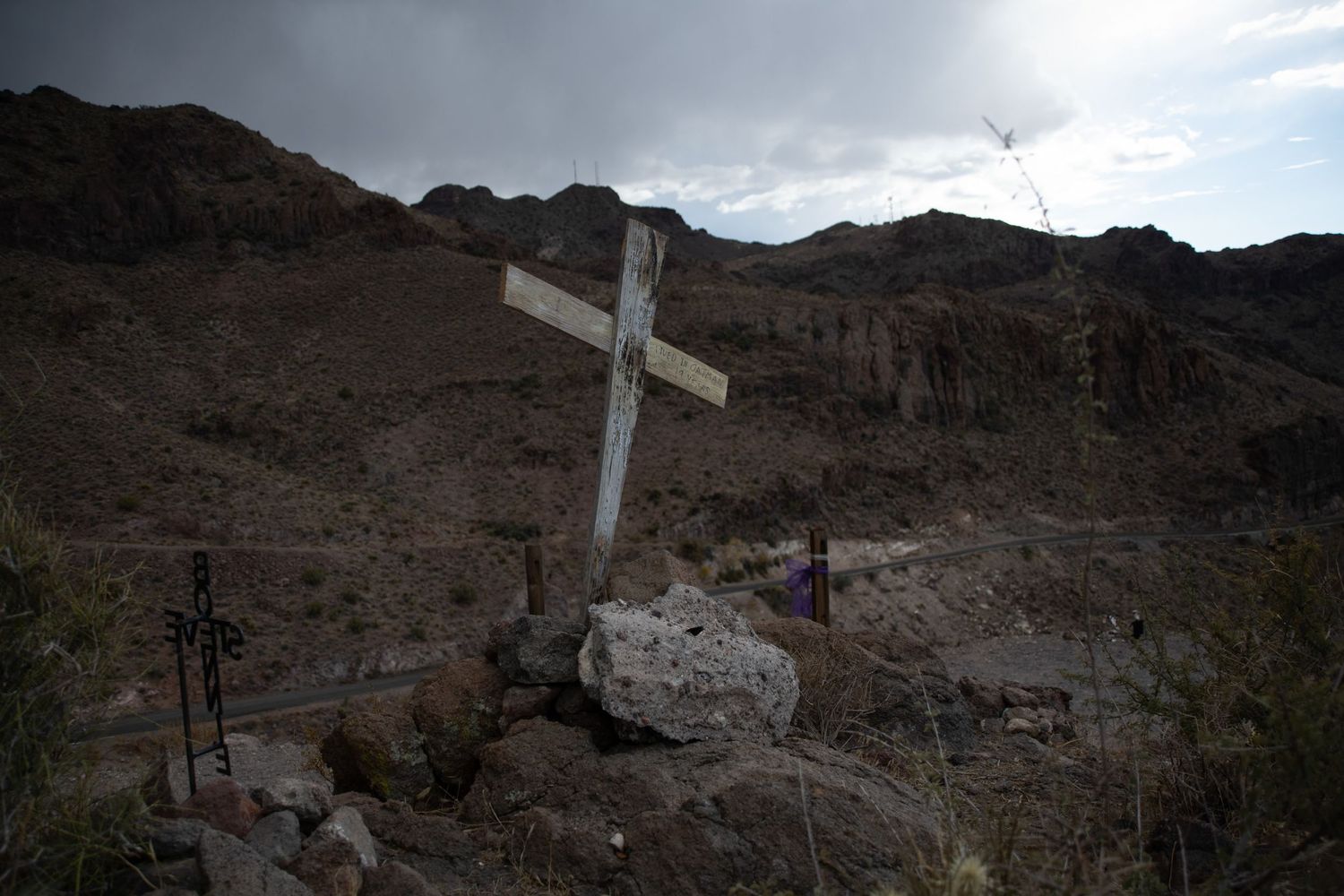 White wooden cross memorial surrounded by rocks in a dramatic desert canyon landscape under cloudy skies.