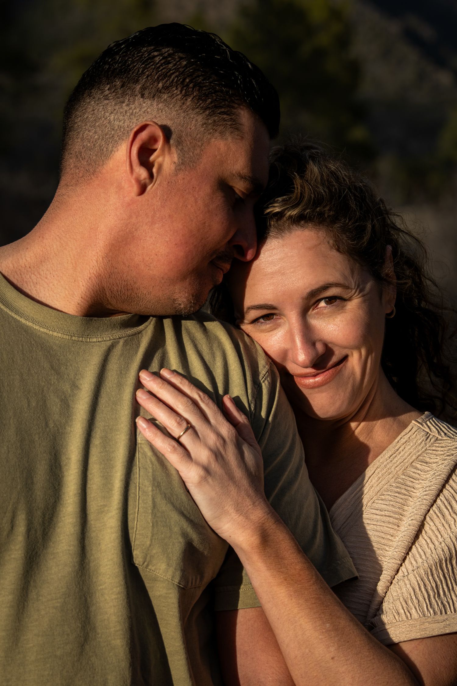 Couple sharing a tender moment outdoors in golden light, woman smiling at camera while resting hand on mans chest.