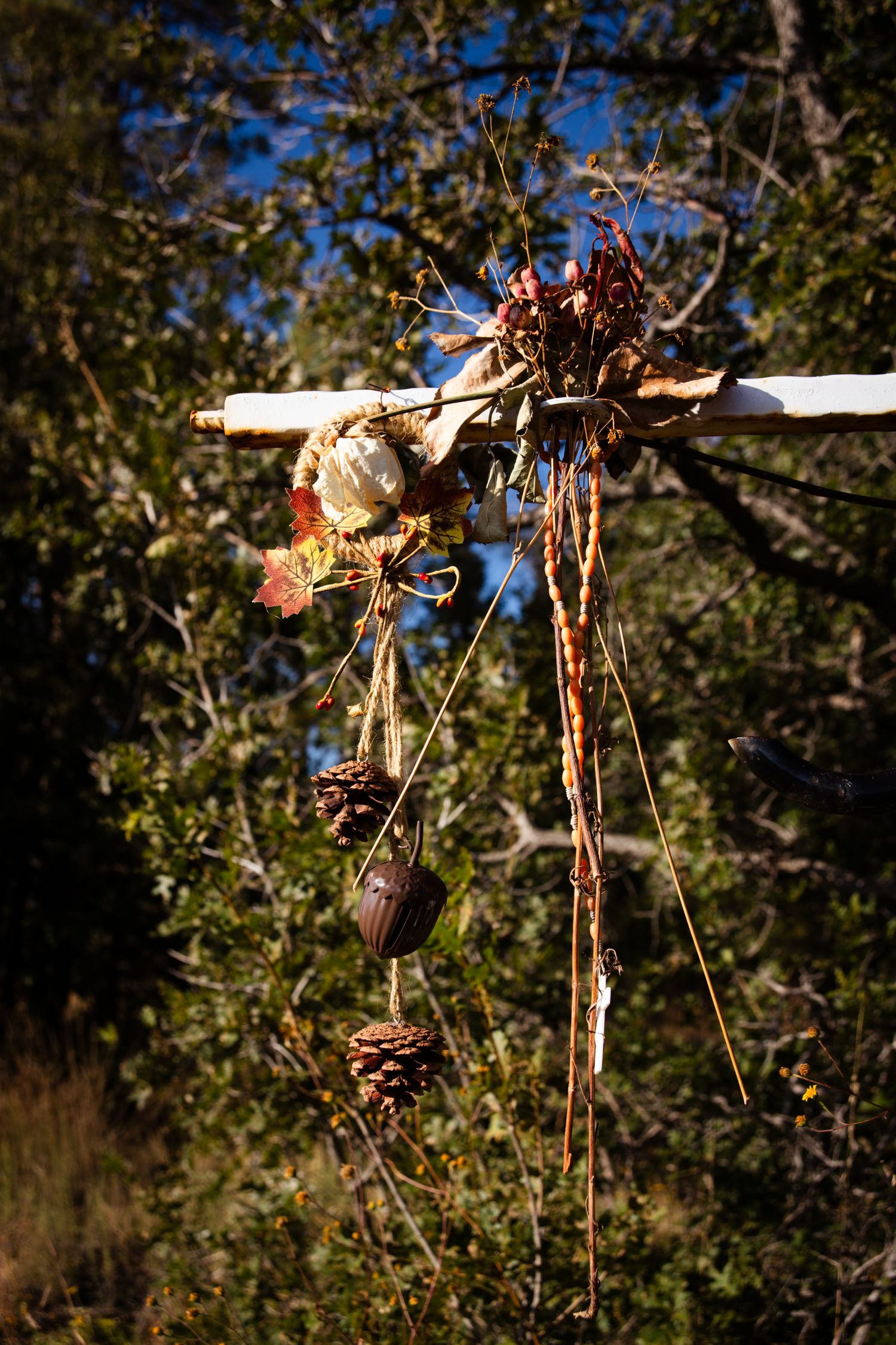 Rustic wooden cross decorated with dried flowers, pine cones, and natural elements hanging outdoors.
