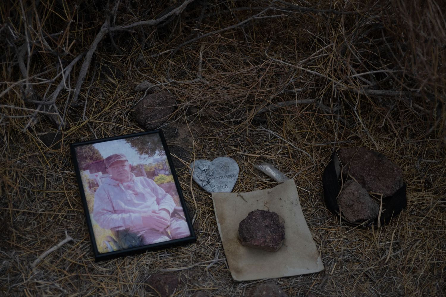 Outdoor memorial with a framed photograph, heart-shaped stone, and small objects on dry desert brush.