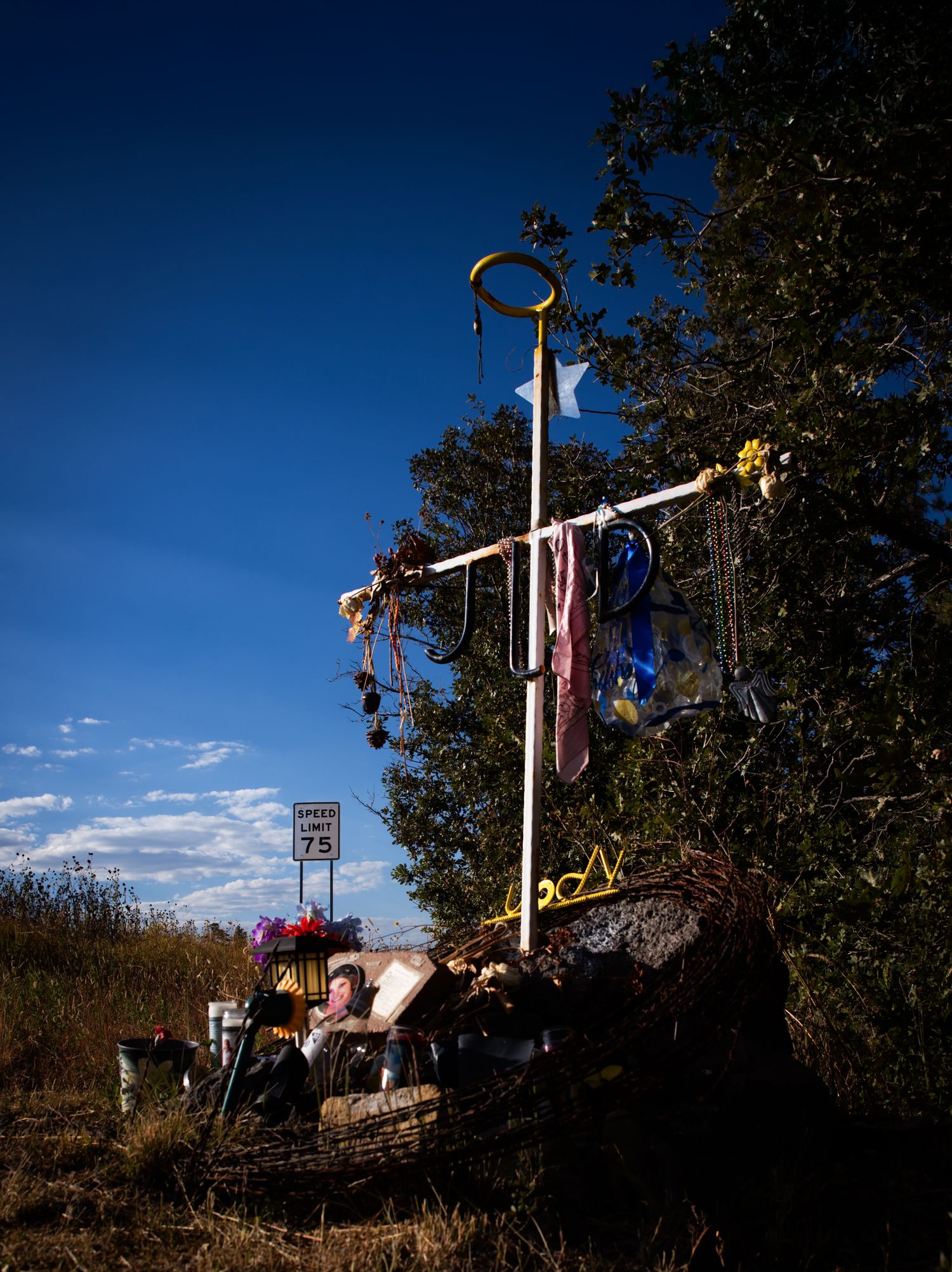 A decorated roadside memorial cross adorned with colorful items near a speed limit sign.