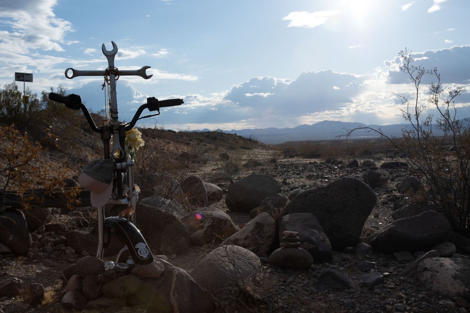 Desert roadside memorial cross made of wrenches surrounded by rocks with mountain landscape backdrop.