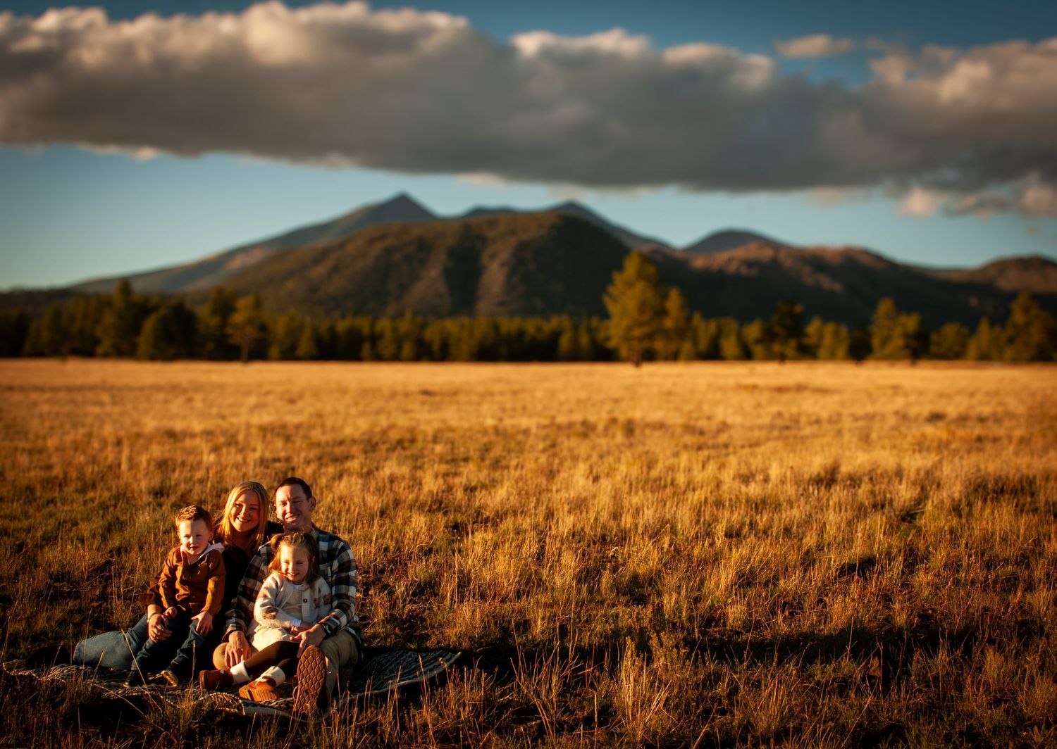 Four children sitting together in golden meadow with mountain range and dramatic clouds at sunset.