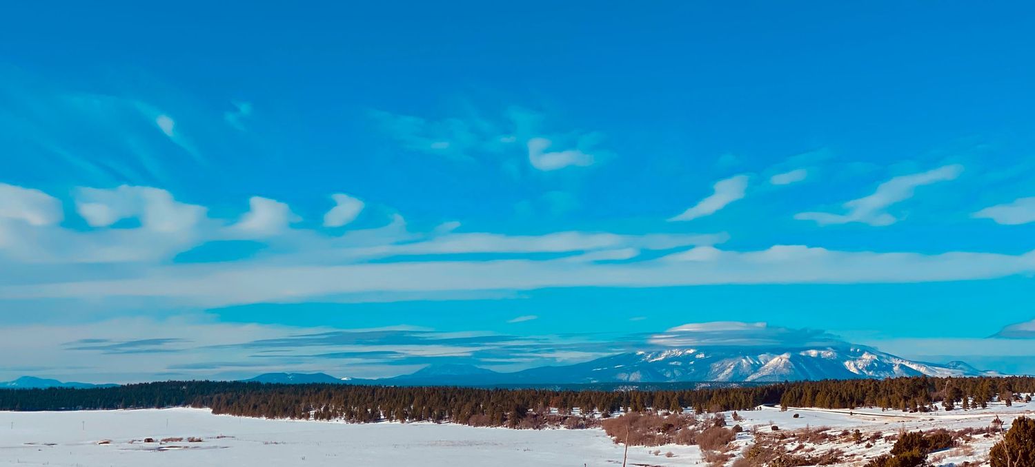 Snow-covered winter landscape with a distant mountain range beneath a vivid blue sky with wispy white clouds.