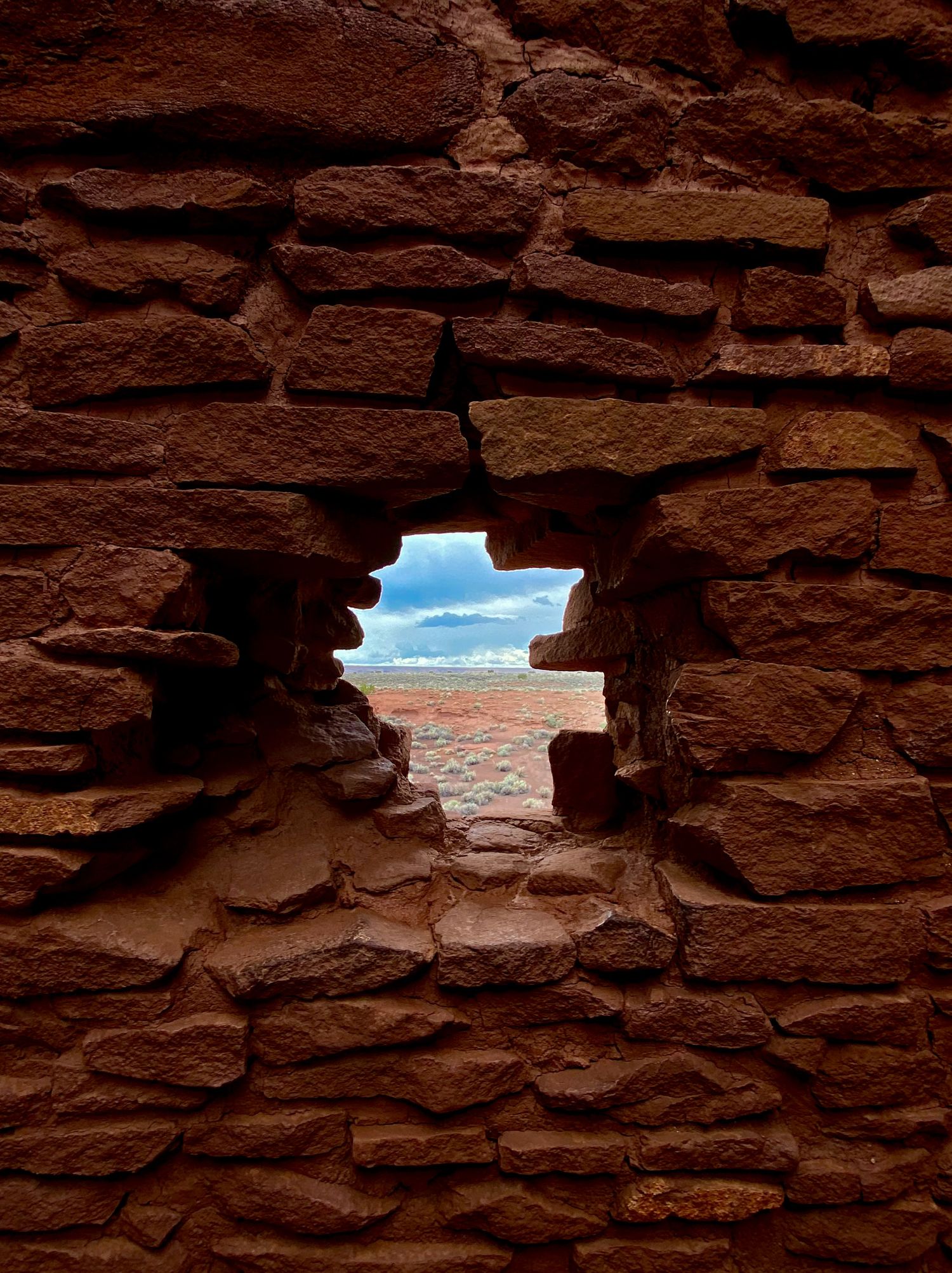 Ancient red sandstone brick wall with a hole revealing a vast desert landscape and cloudy sky.