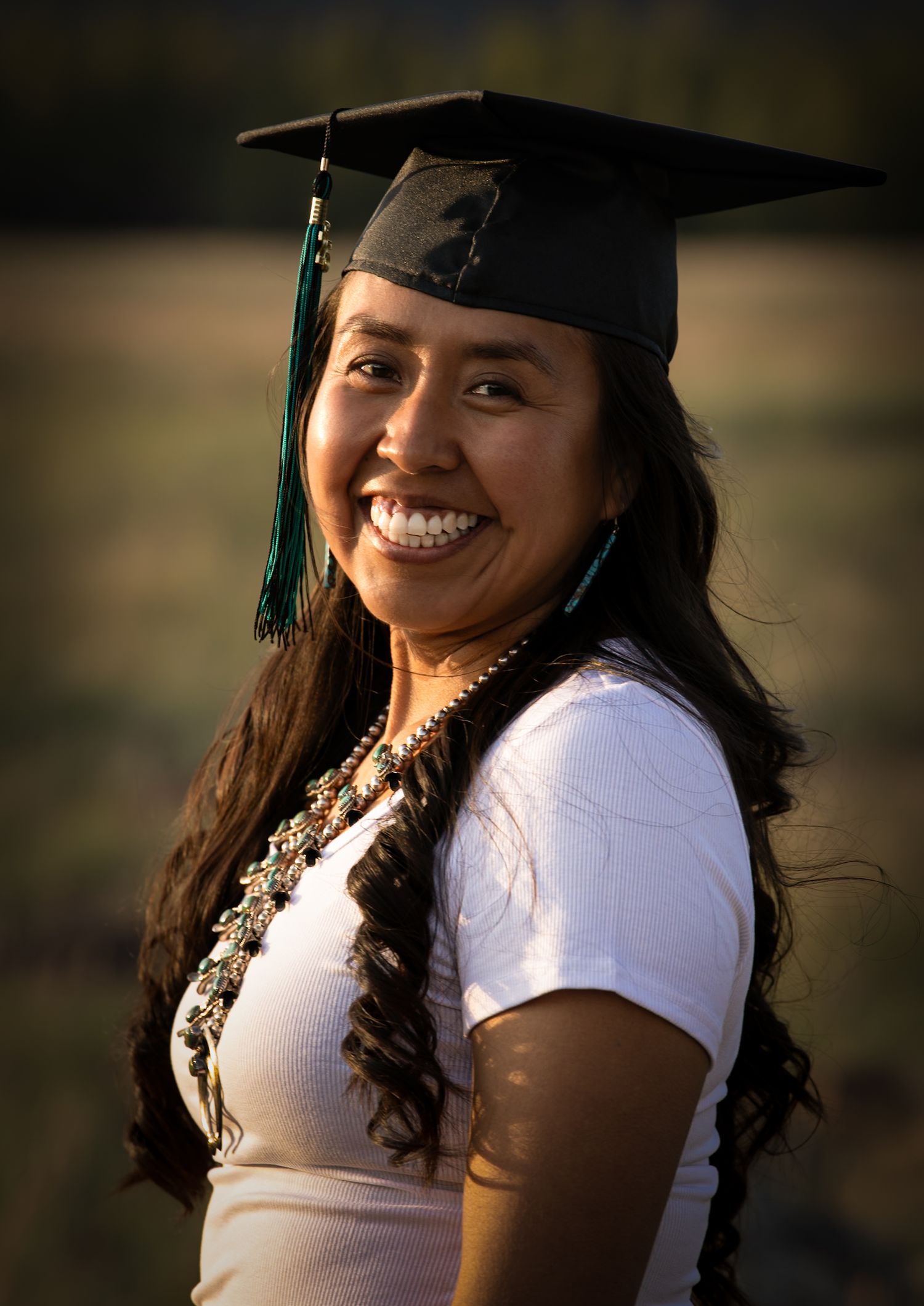 Smiling young woman wearing a graduation cap and traditional Native American jewelry outdoors.