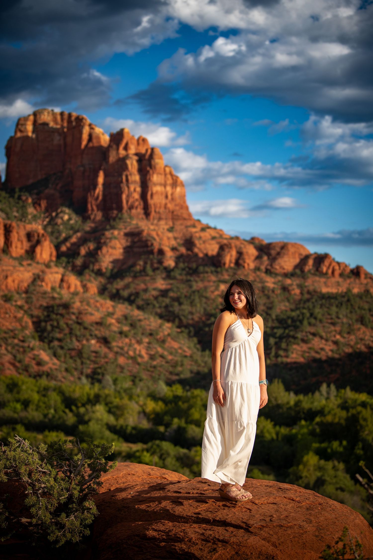 Woman in white dress standing on red rock with Cathedral Rock formation and blue sky in Sedona, AZ.