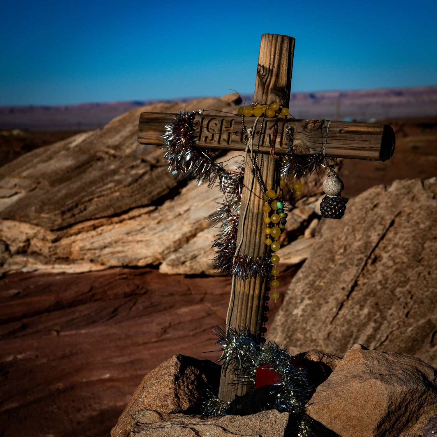 Weathered wooden memorial cross adorned with greenery and beads on red rock desert landscape.