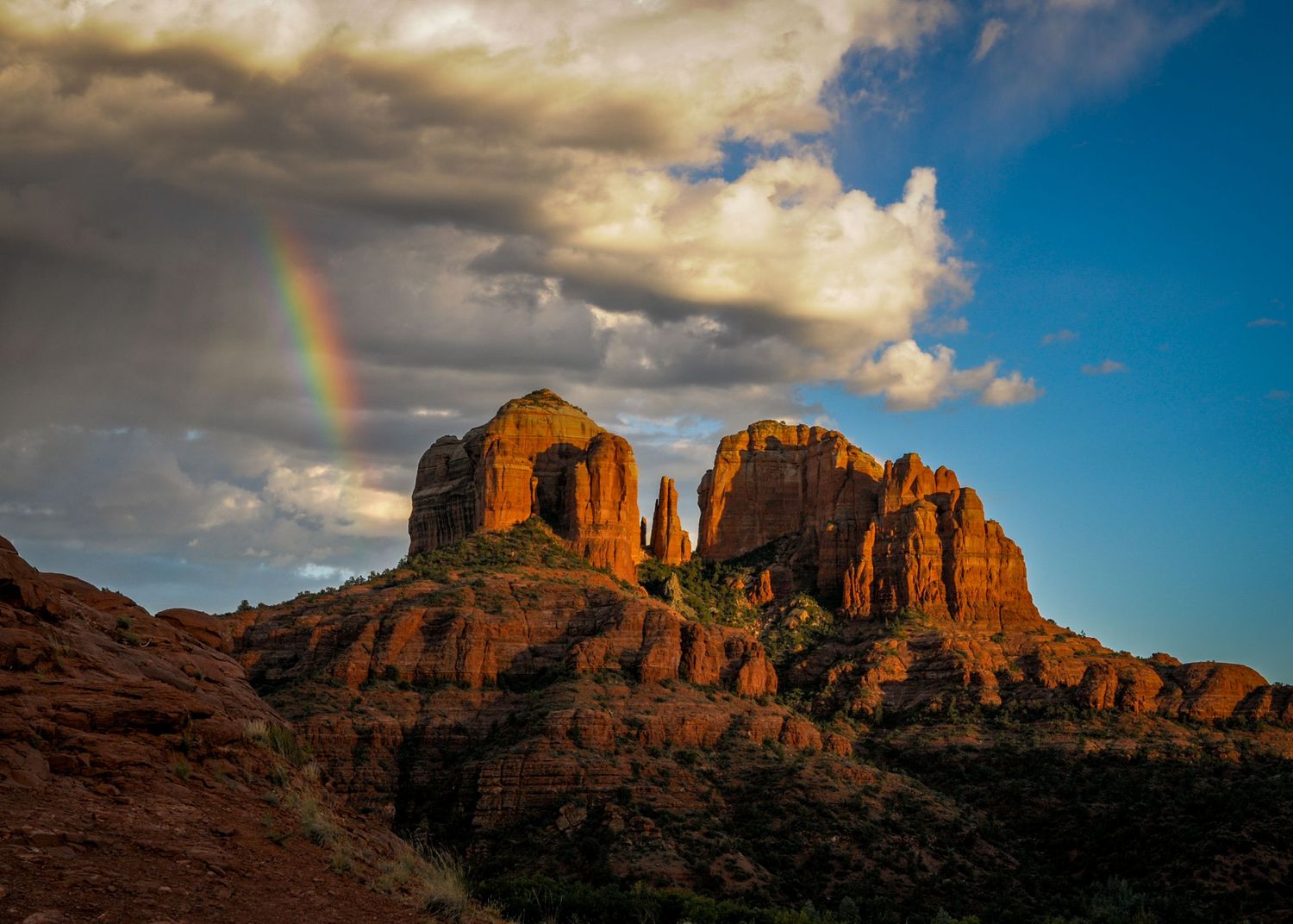Red rock formations of Sedona, Arizona glow under dramatic storm clouds with a vibrant rainbow arching across the blue sky.