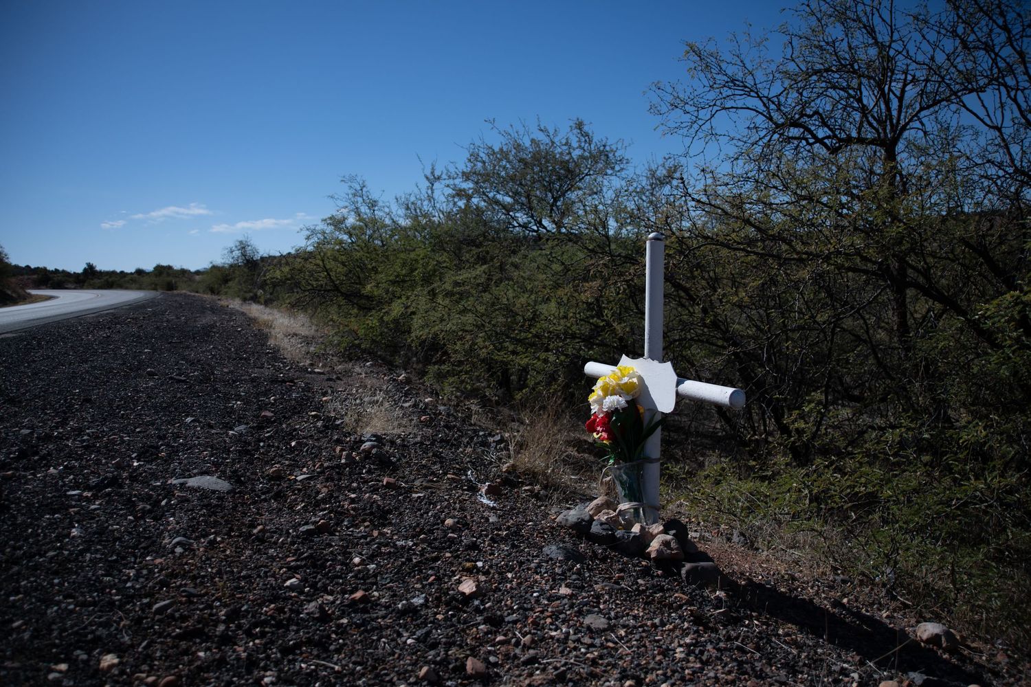 Solitary white memorial cross with flowers standing on gravel beside a desert road.