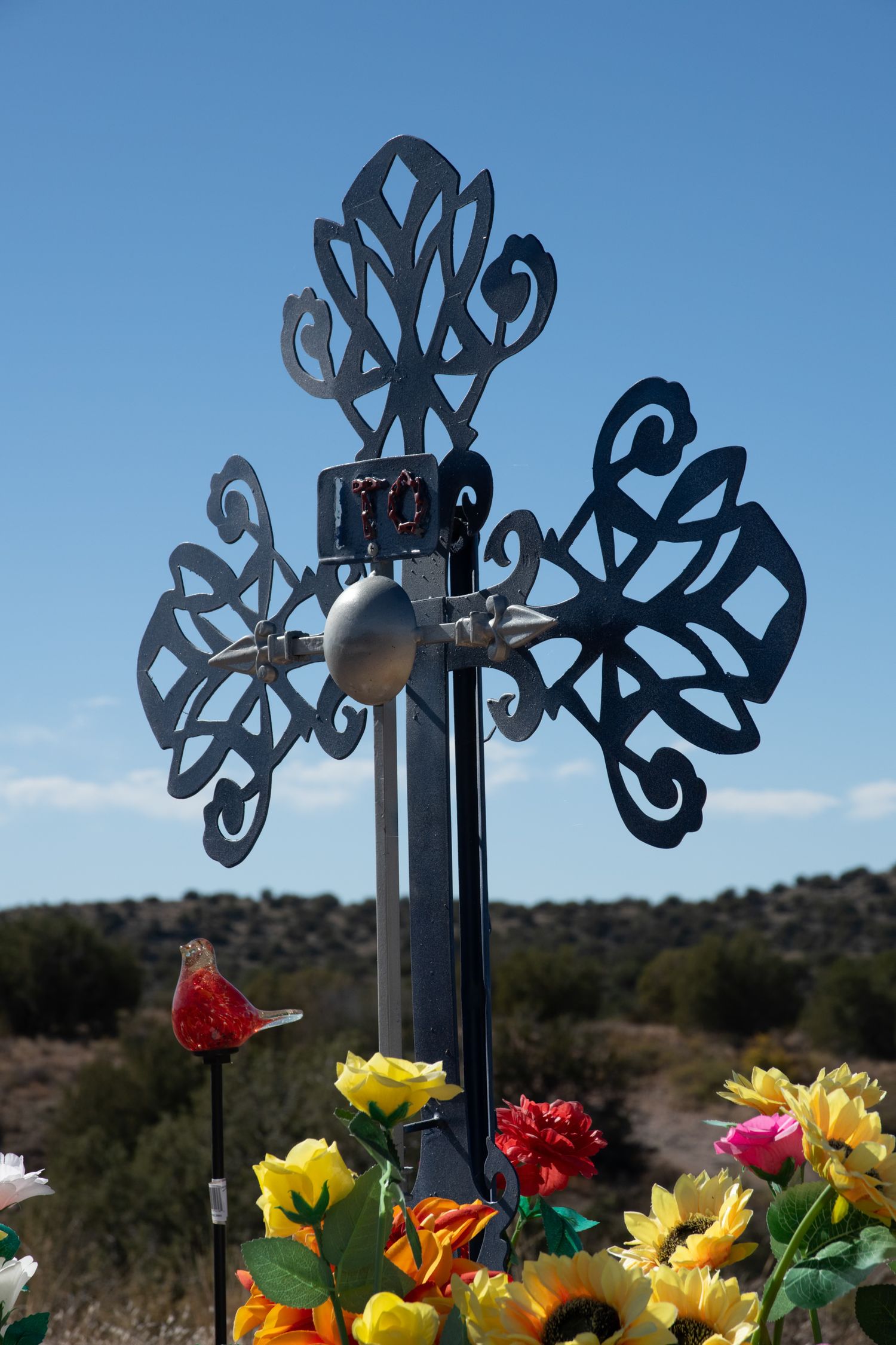 Decorative black metal cross roadside memorial with colorful artificial flowers in a desert setting.