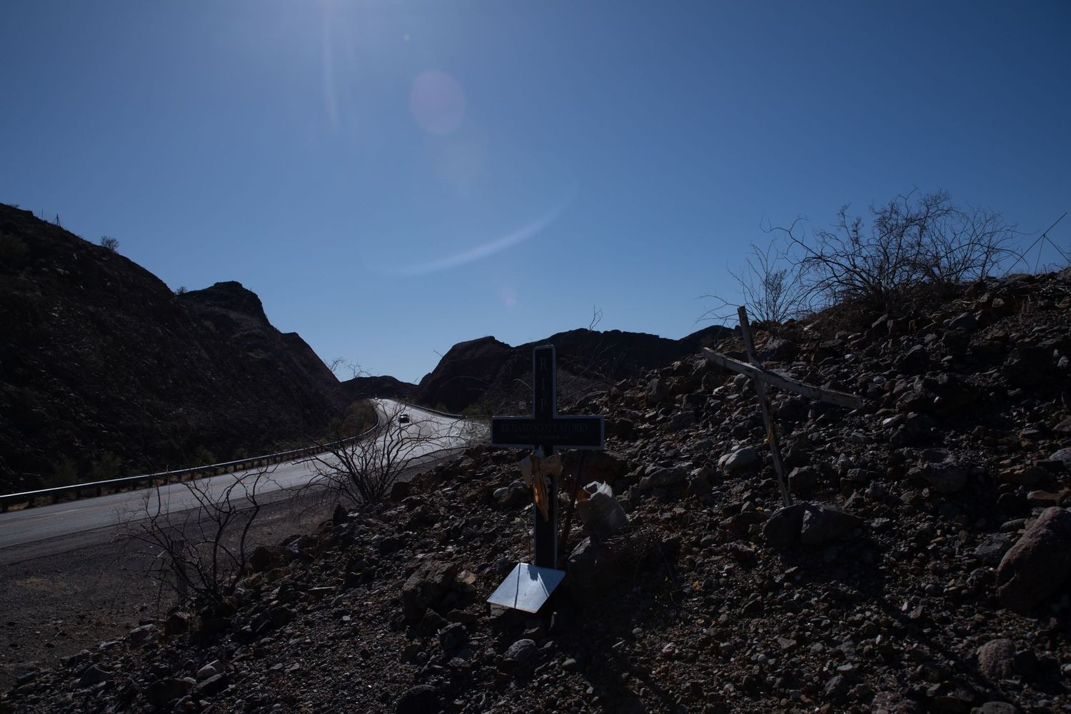 A dark roadside memorial cross stands on rocky desert hillside under bright blue sky.