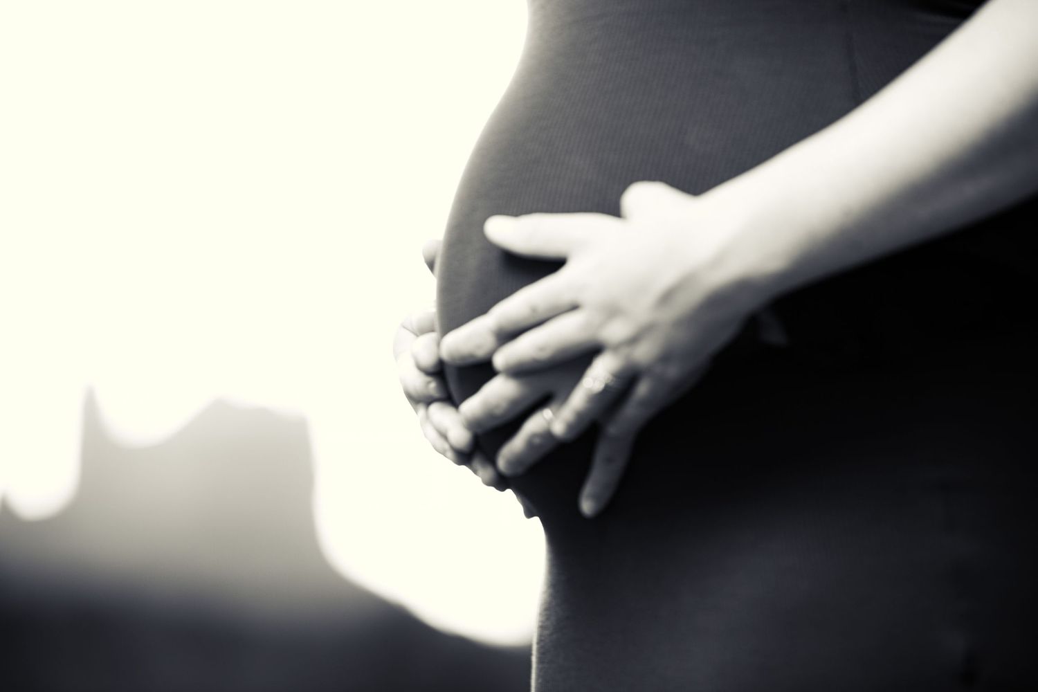 Black and white close-up of a pregnant woman cradling her baby bump with both hands outdoors.