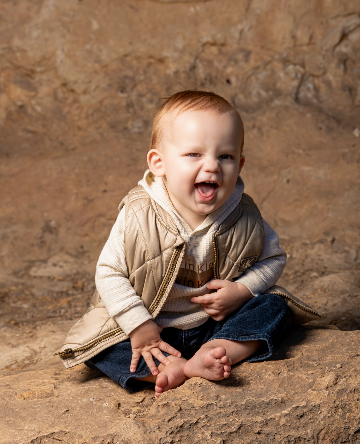 Happy red-haired baby sitting on sandy ground, laughing joyfully, wearing a beige vest and jeans.
