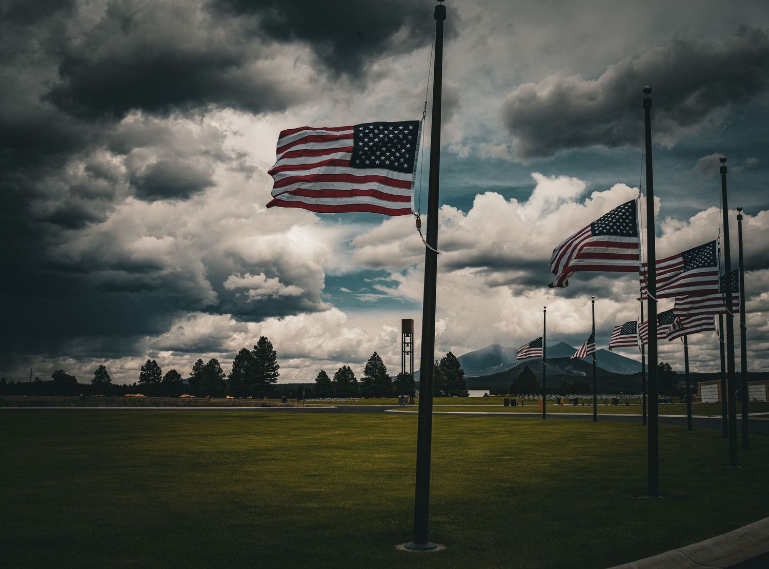 Multiple American flags on tall poles waving under dramatic stormy cloudy skies over green field.