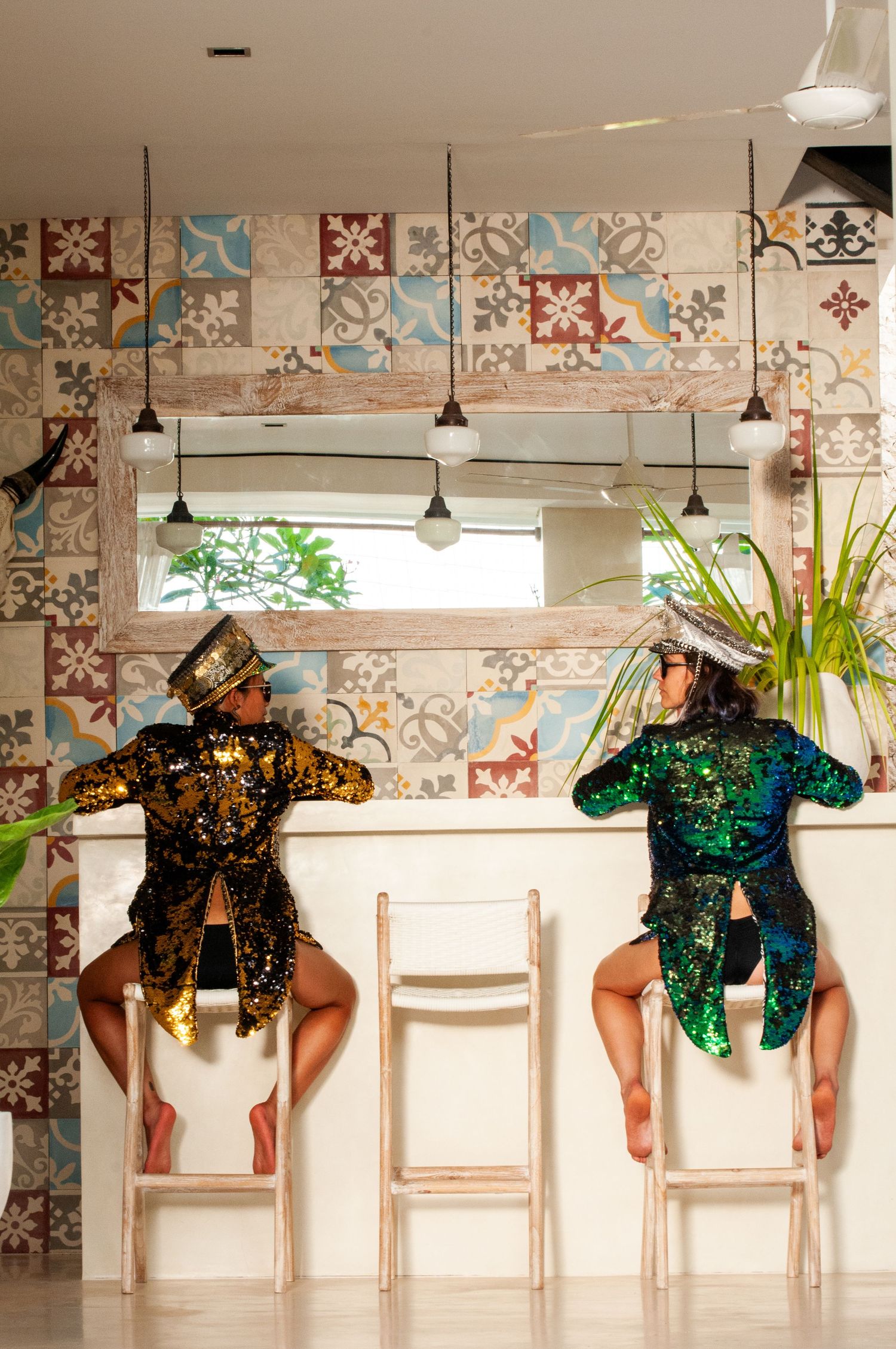 Two performers in sequined costumes and top hats sitting at a bar with decorative tiled walls.
