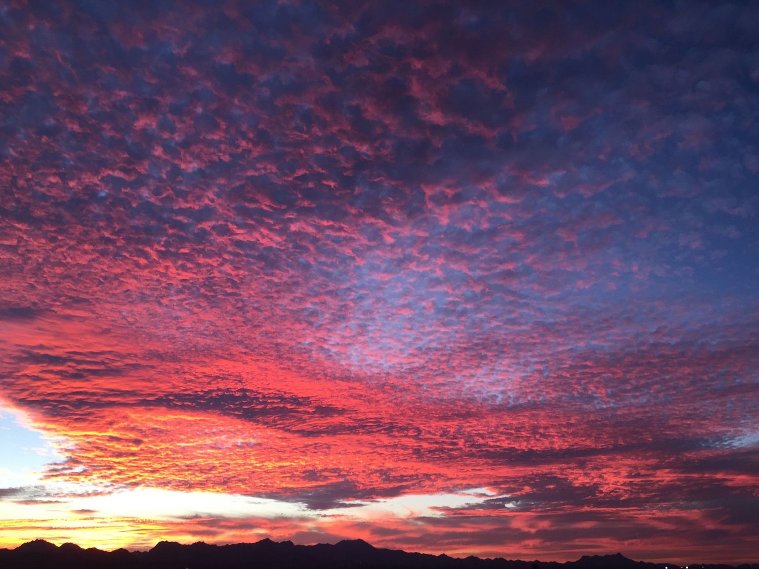Vivid red and pink sunset sky with dramatic cloud formations over a mountain silhouette.