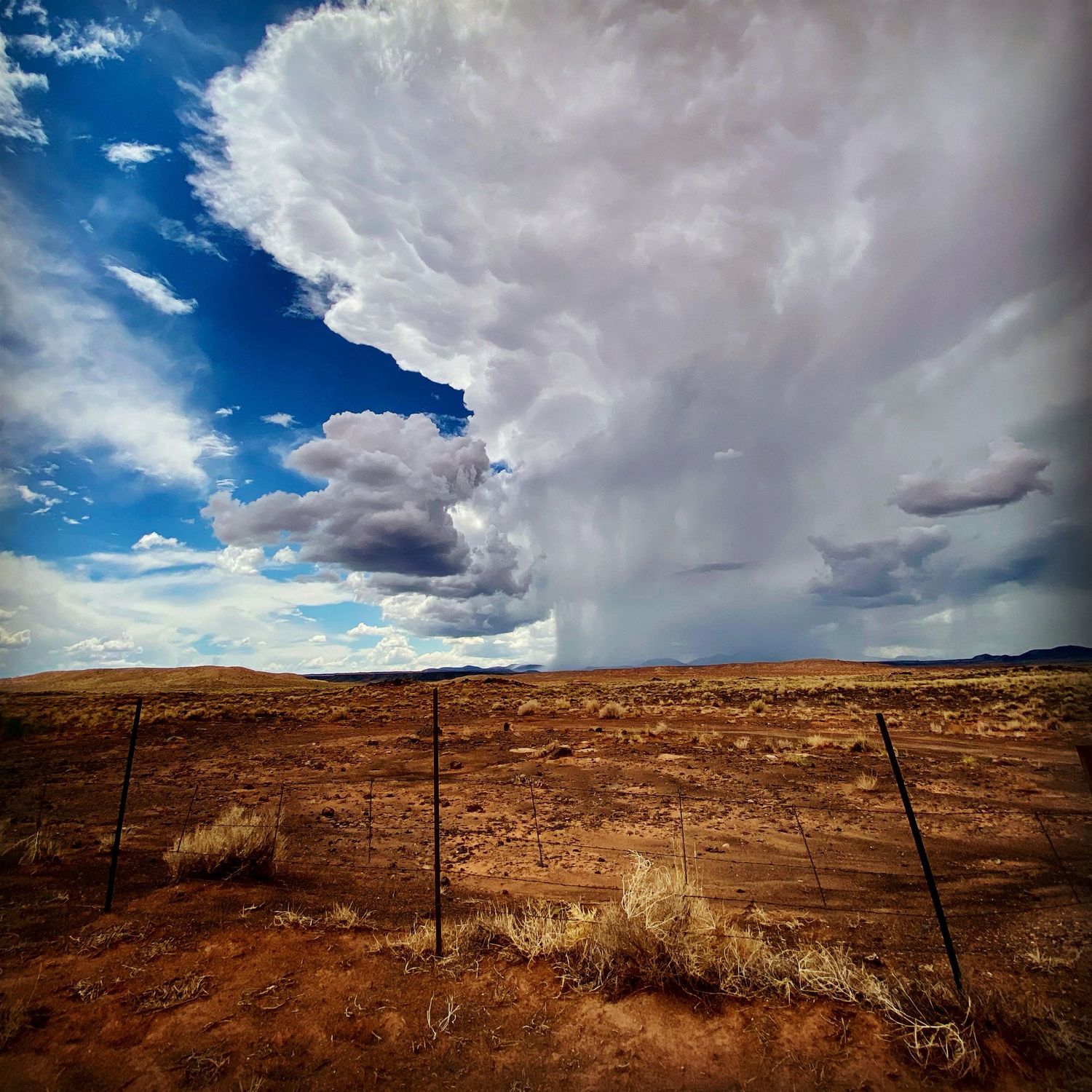 Dramatic storm clouds building over arid desert landscape with rustic fence in foreground.