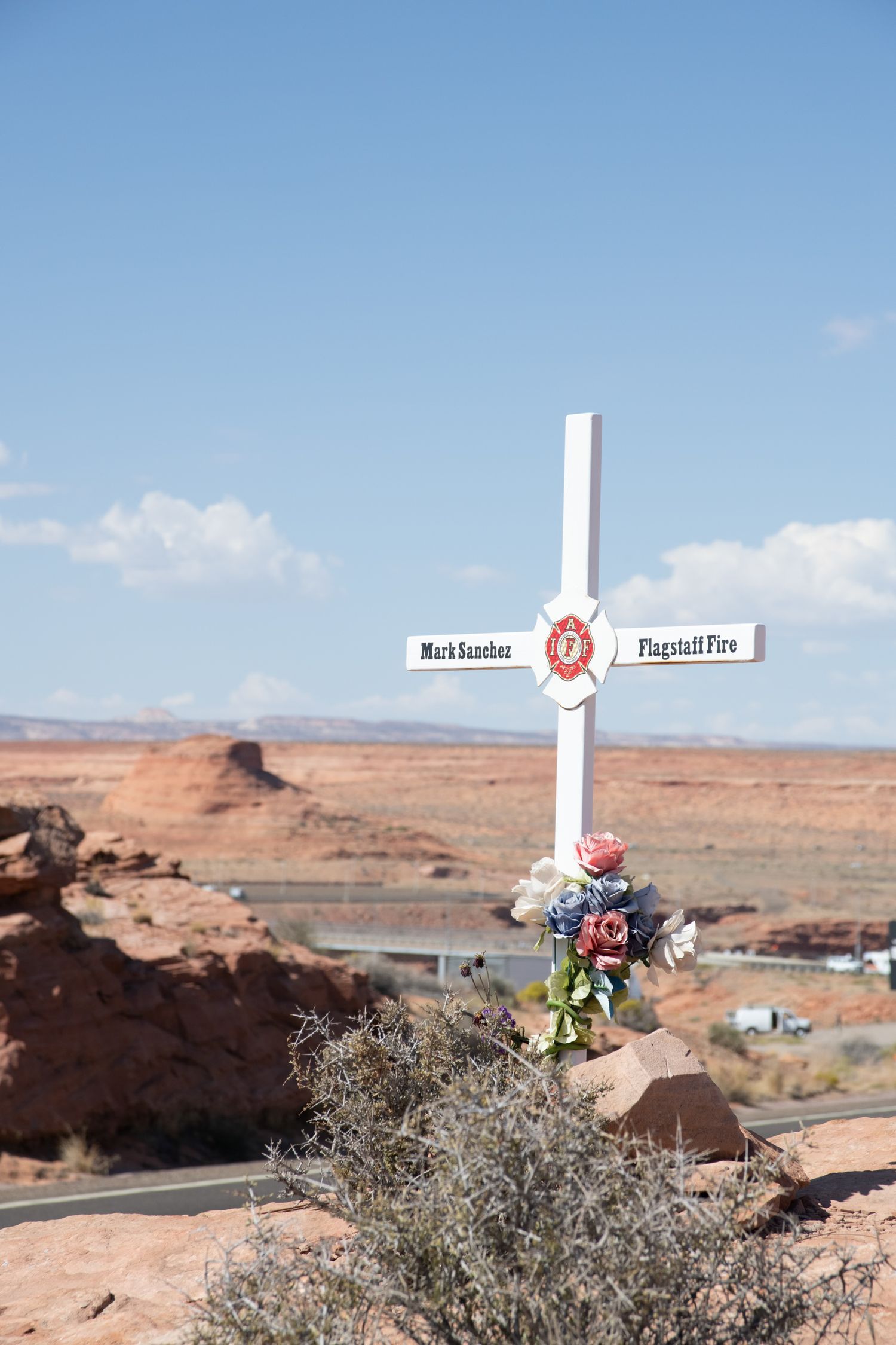 White memorial cross for Mark Sanchez of Flagstaff Fire decorated with flowers in Arizona desert landscape.