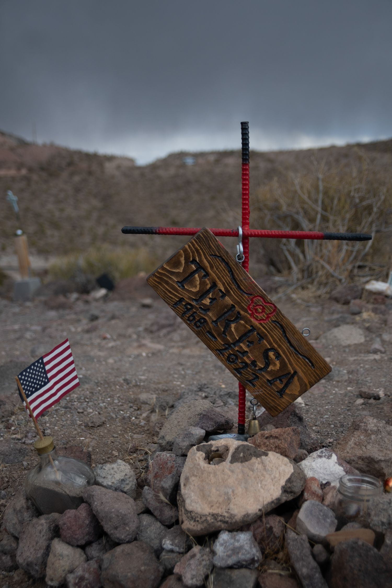 A rustic wooden cross memorial with an American flag marks a desert grave site under stormy skies.