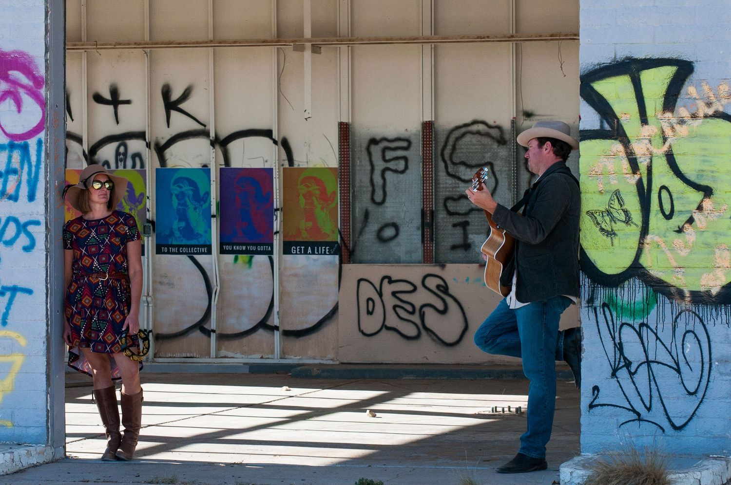 Woman in floral dress and man playing guitar posing in front of a graffiti-covered urban wall.
